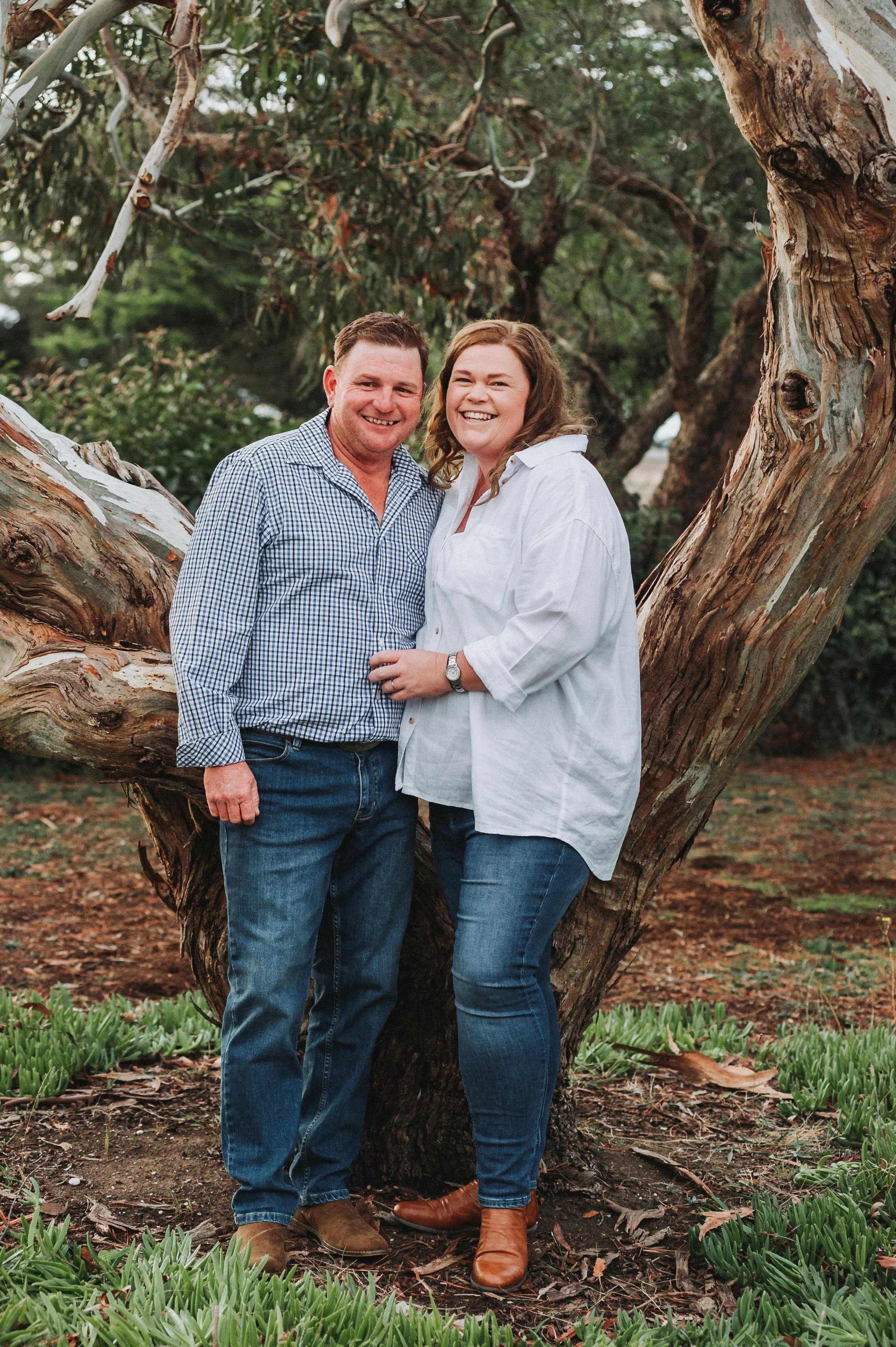 A happy couple standing side by side in front of a large tree, smiling at the camera. They are dressed casually in jeans and shirts, surrounded by greenery.