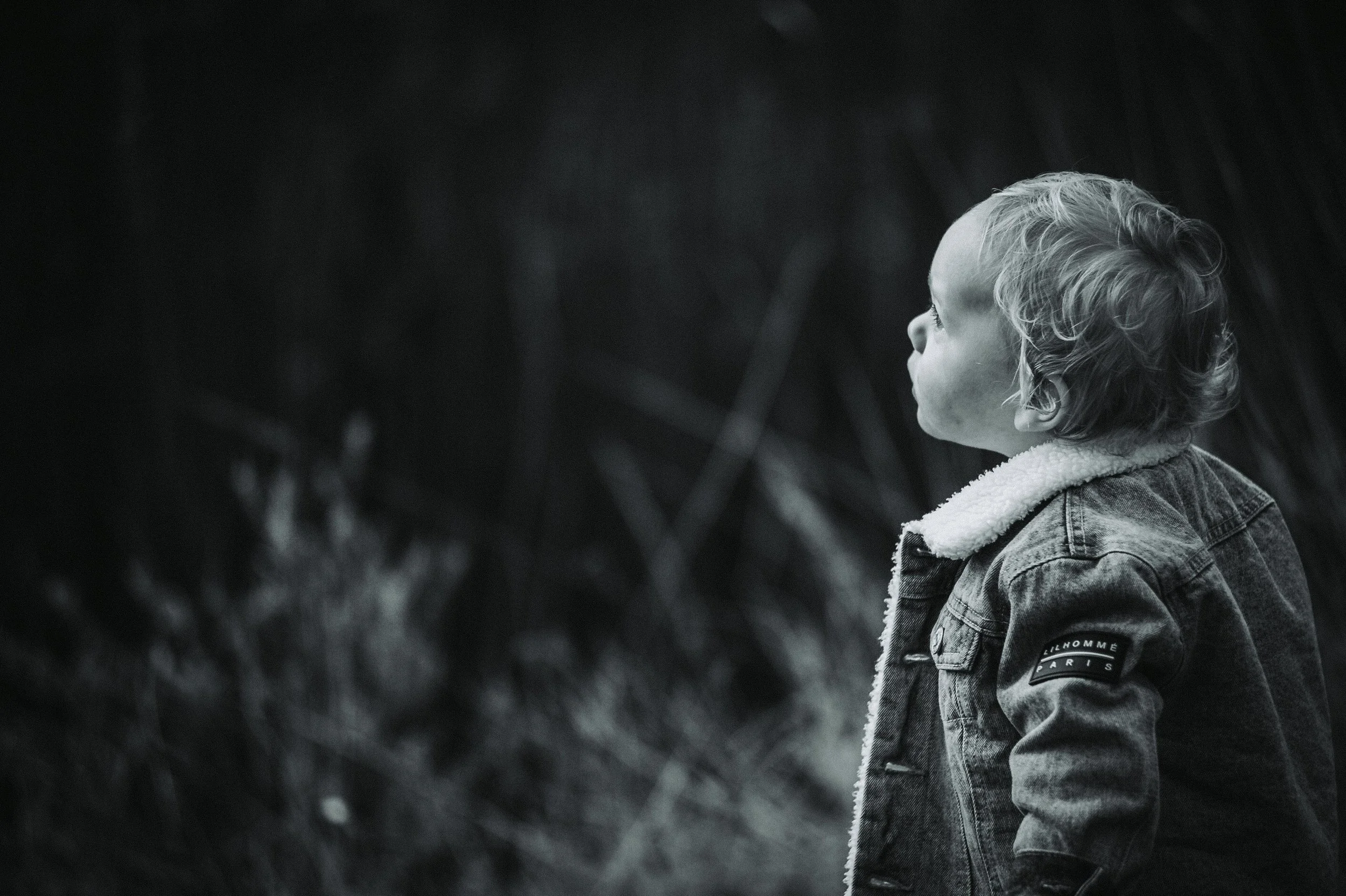Black and white photo of a child in a denim jacket looking upward with a blurred nature background.