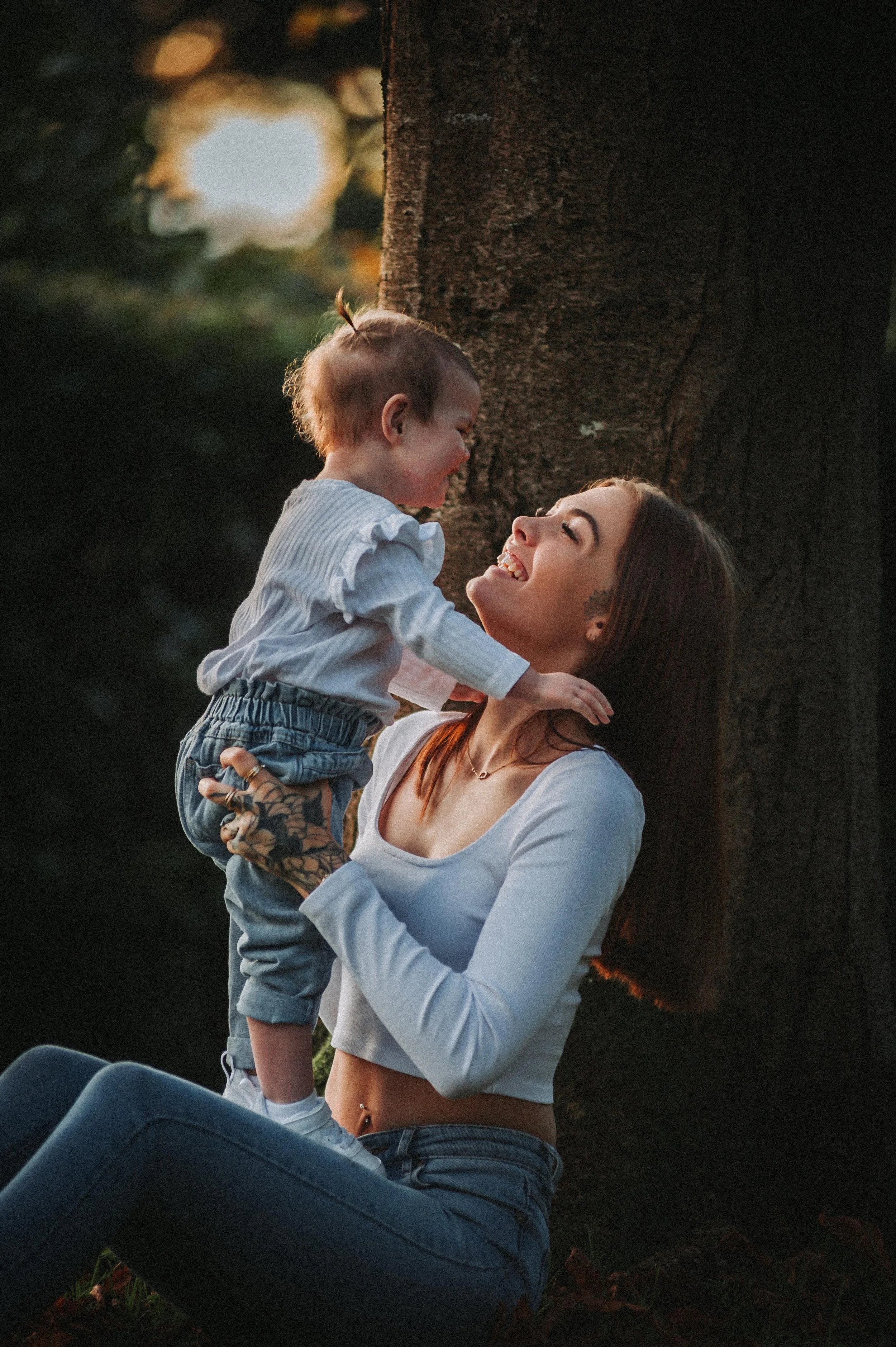 A woman smiling and holding a baby outdoors near a tree.