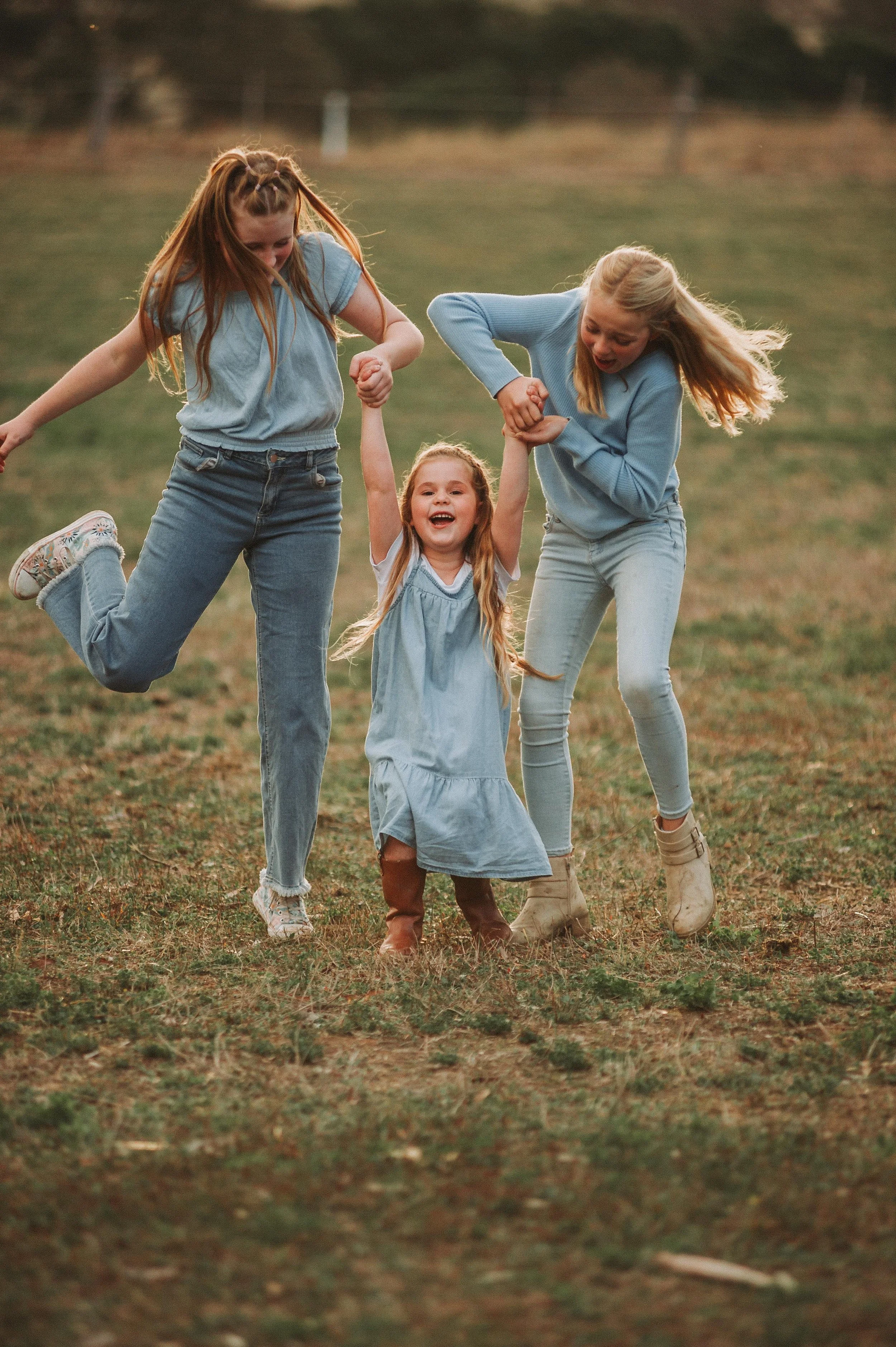 Three girls playing in grass, two older girls holding hands with a younger girl, all wearing blue clothing.