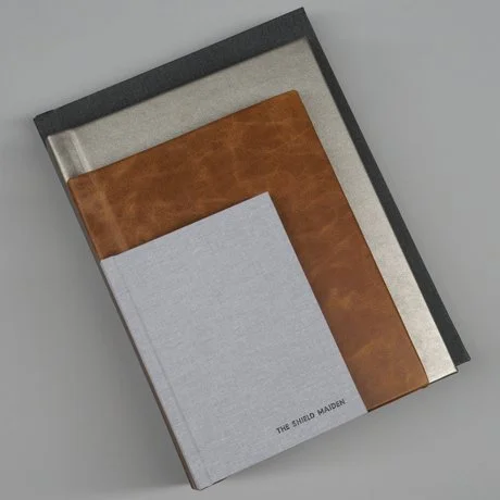 Three hardcover books stacked; one in gray and black, one in brown and silver, and one in light gray titled 'The Shield Maiden.'