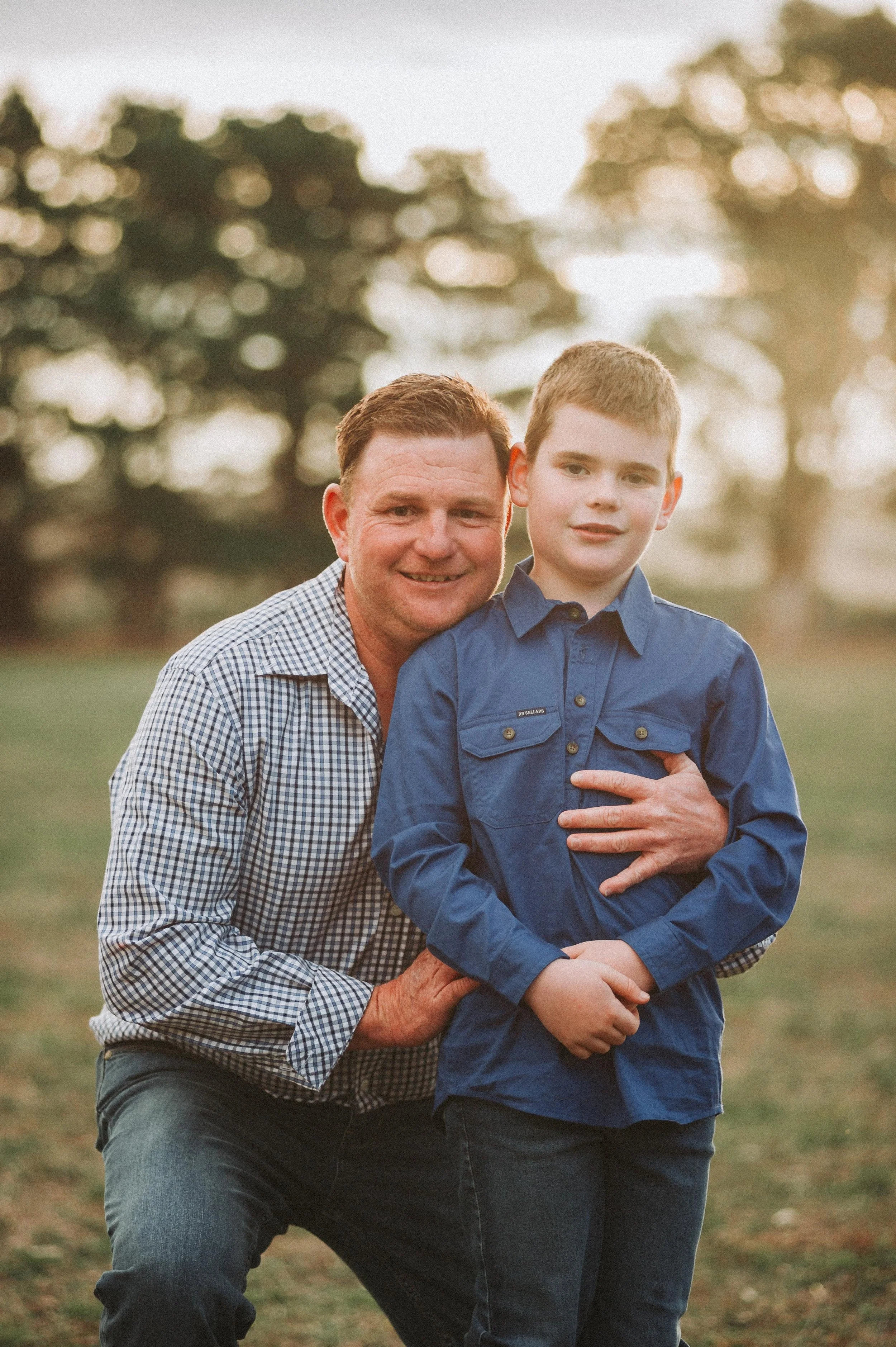 Man hugging a boy in a blue shirt, outdoors with trees and a grassy field in the background