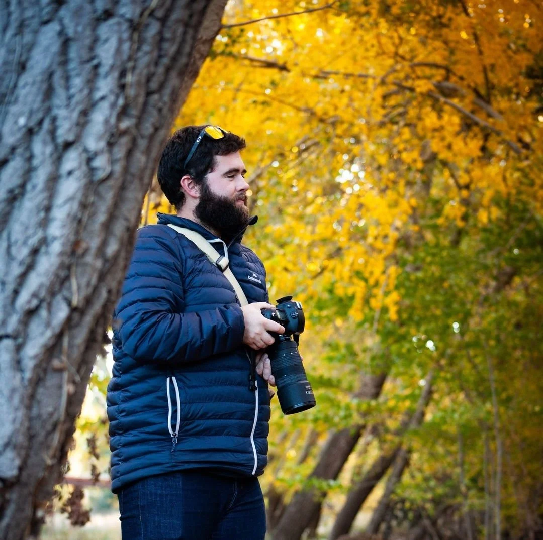 Photographer in a blue jacket holding a camera with a telephoto lens, standing in a forest with autumn foliage.