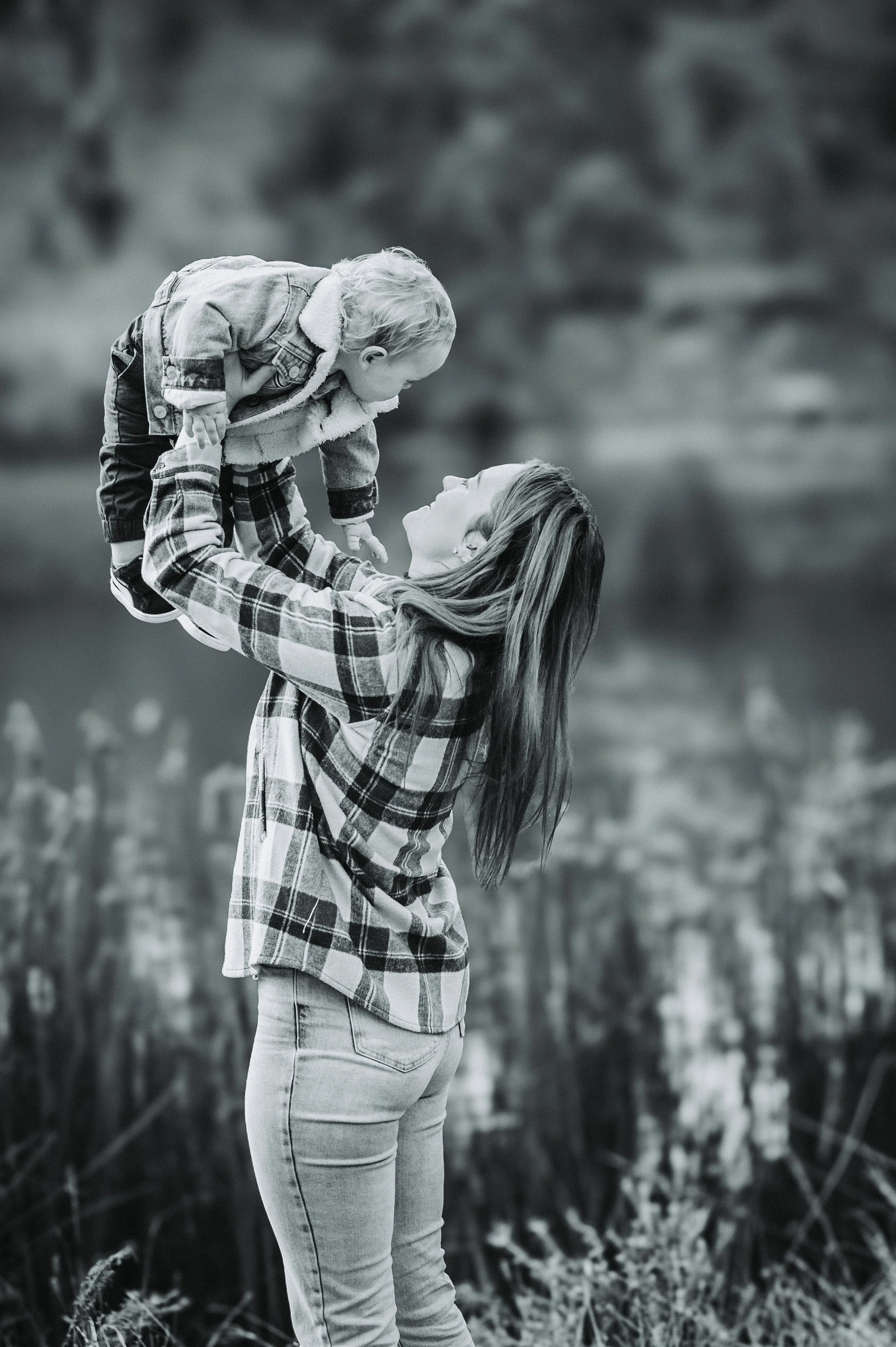 A woman lifting a toddler in the air outdoors, both smiling, with tall grass in the background.