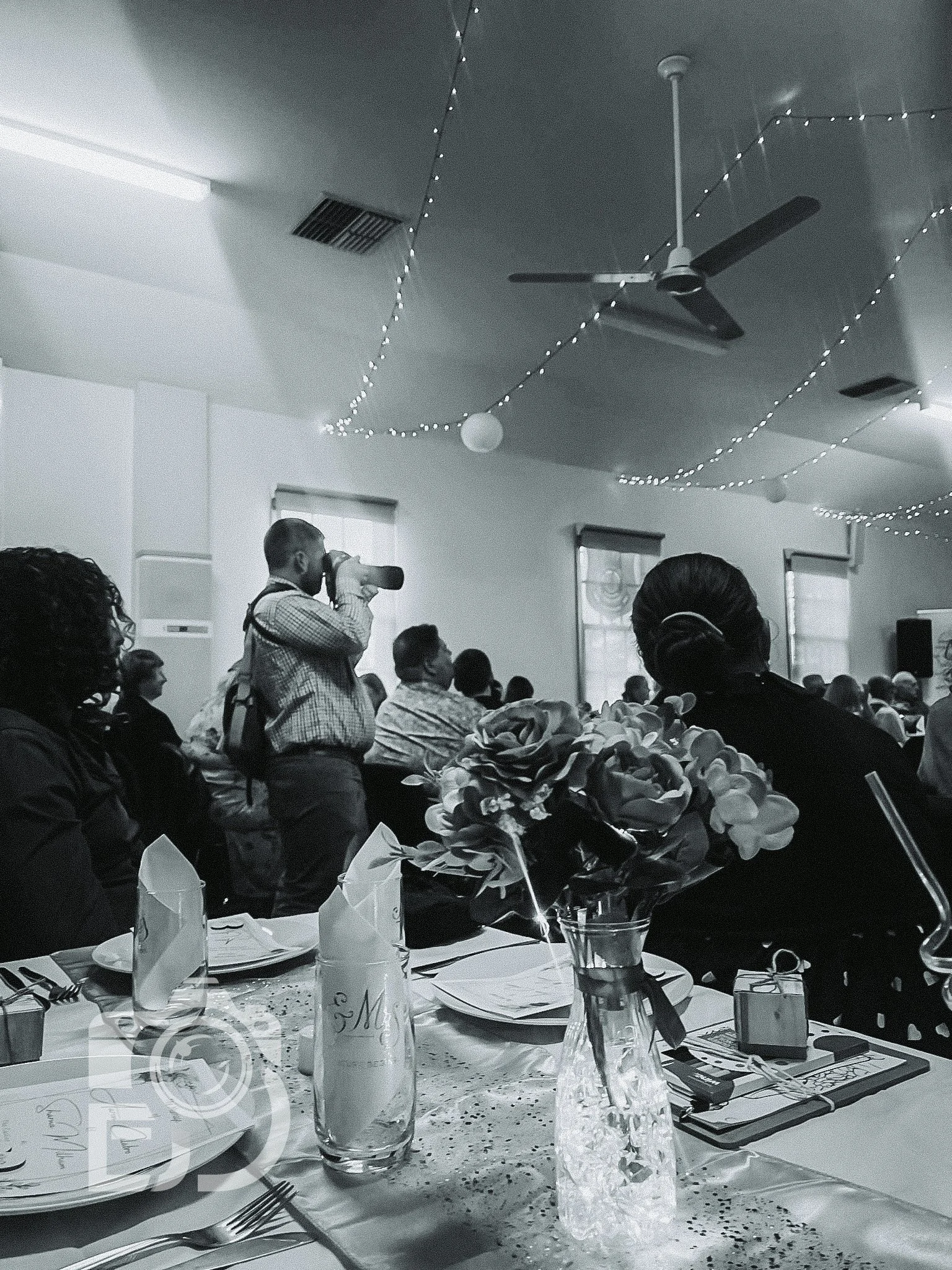 Indoor event with a photographer taking pictures, tables set with plates and glasses, floral centerpiece, and decorative lighting.
