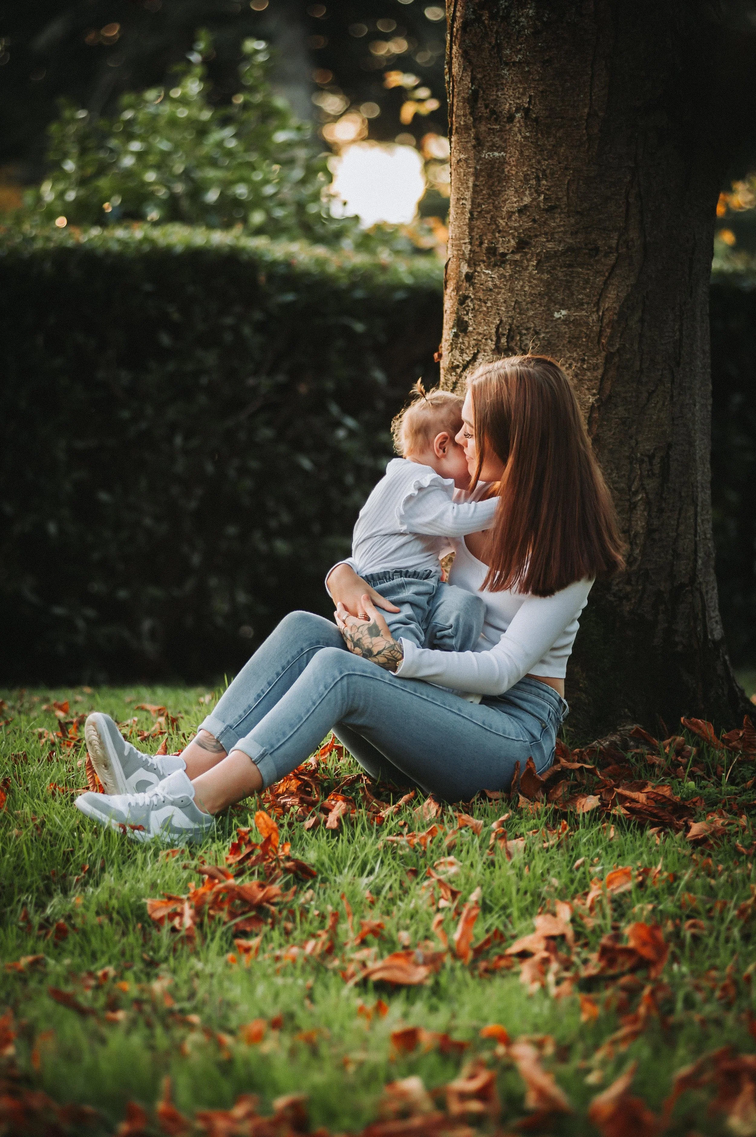 Woman sitting under a tree holding a baby, surrounded by autumn leaves on the grass.