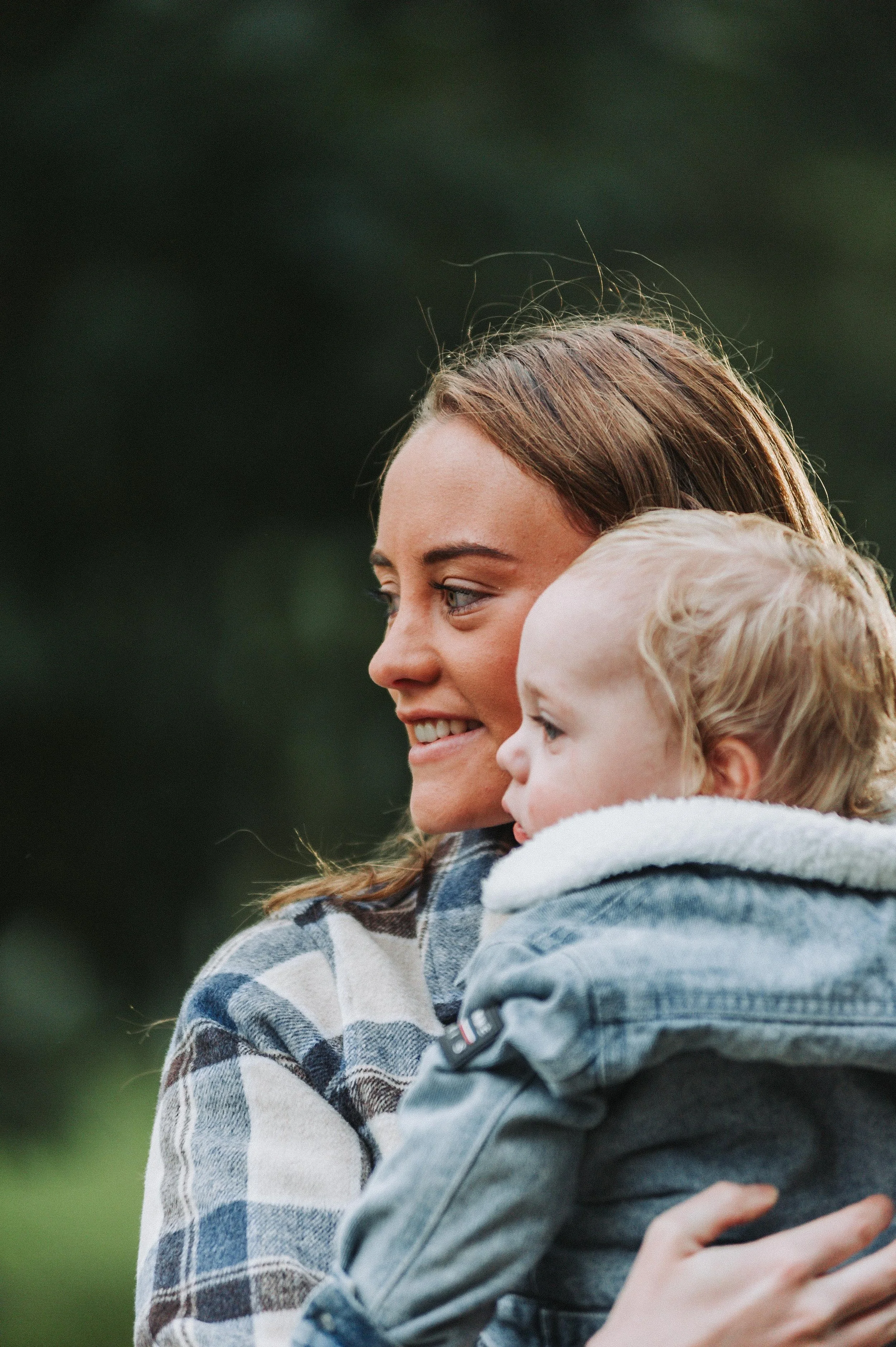 Woman holding a child outdoors, both wearing jackets, with a blurred green background.