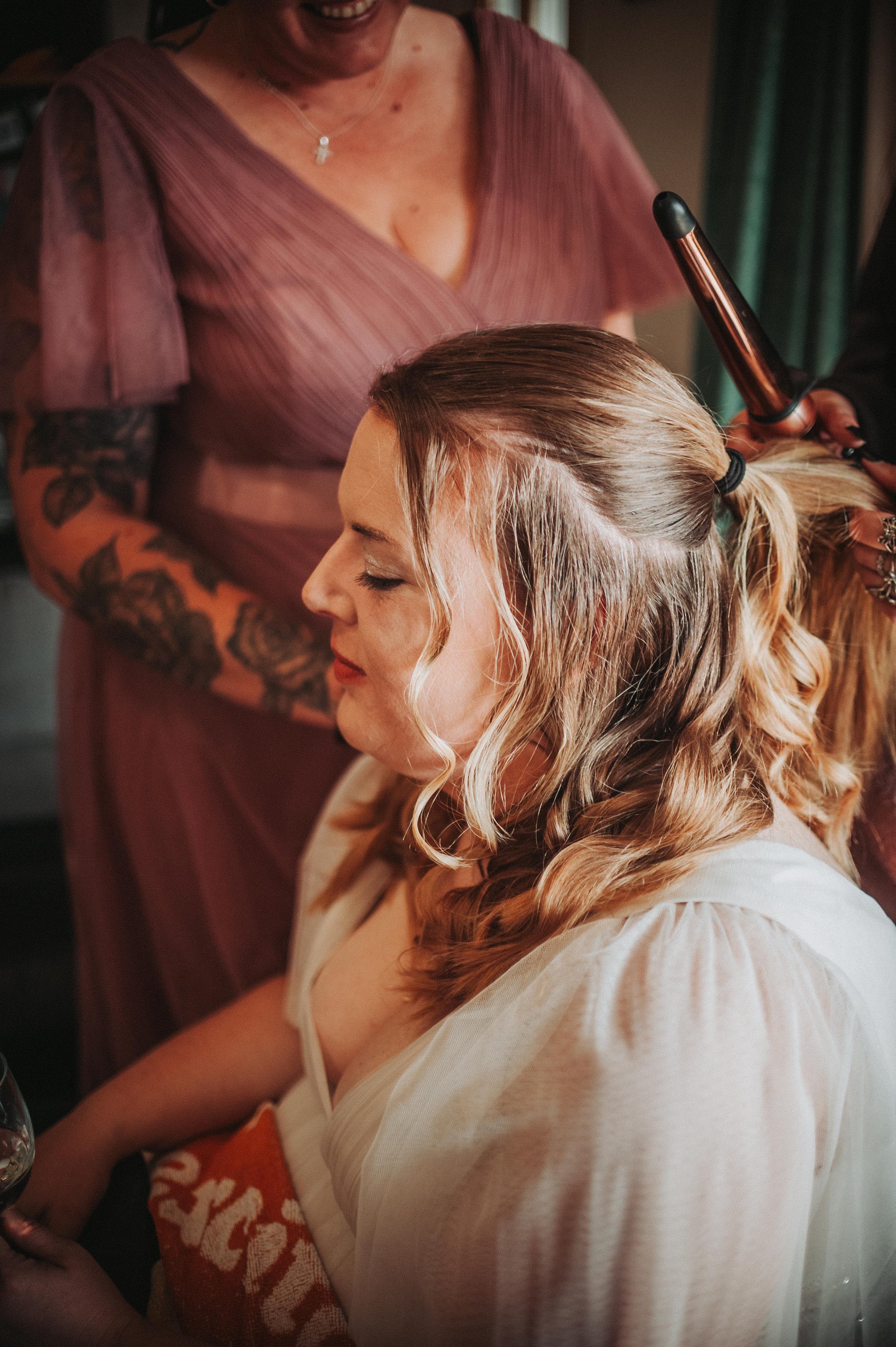 Woman having her hair styled with a curling iron, wearing a white robe, seated indoors. Another person with tattoos, wearing a mauve dress, assists with the hairstyling.