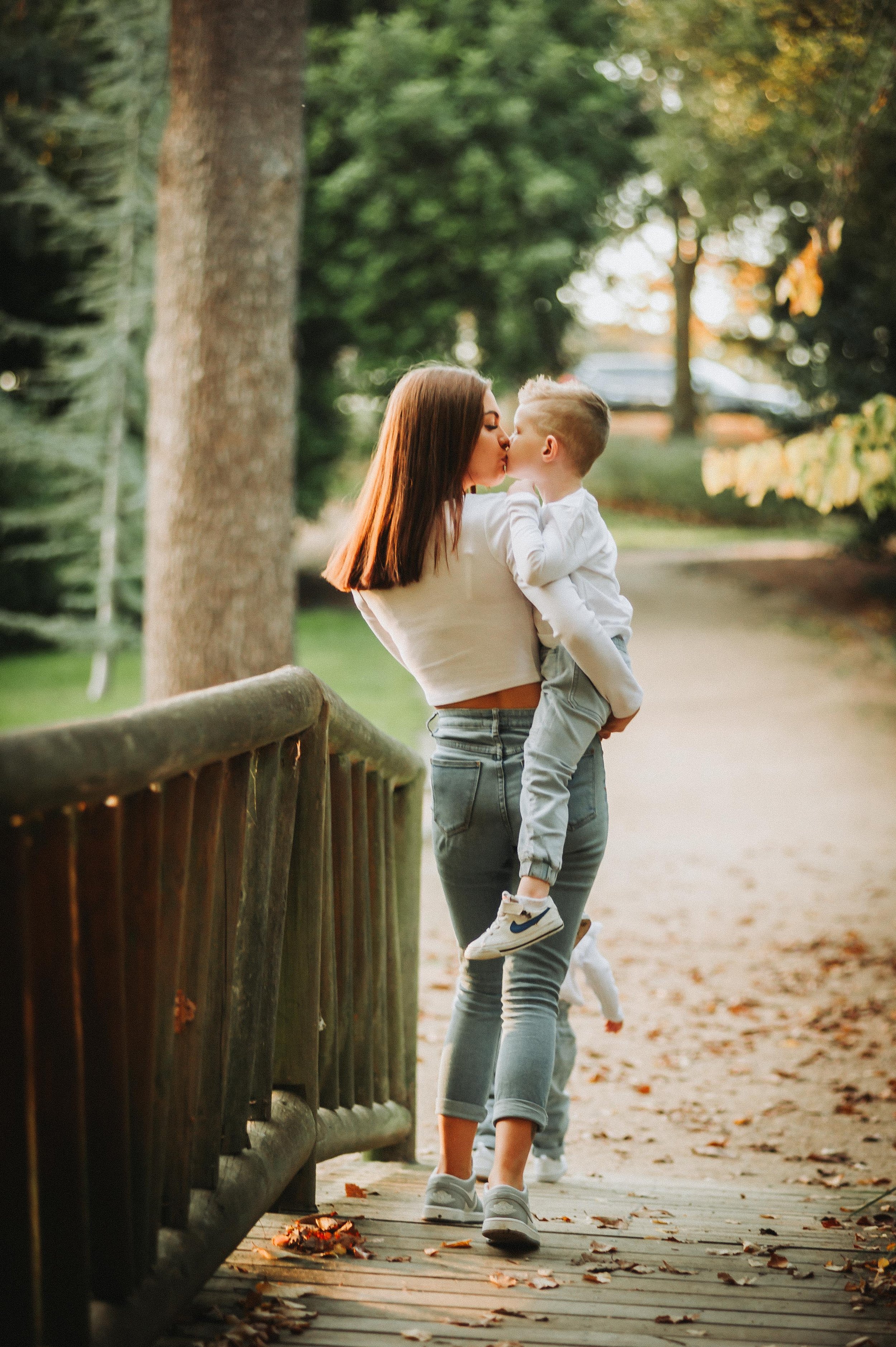 A woman holding and kissing a child on the cheek while standing on a wooden bridge in a park, with trees and a path in the background.