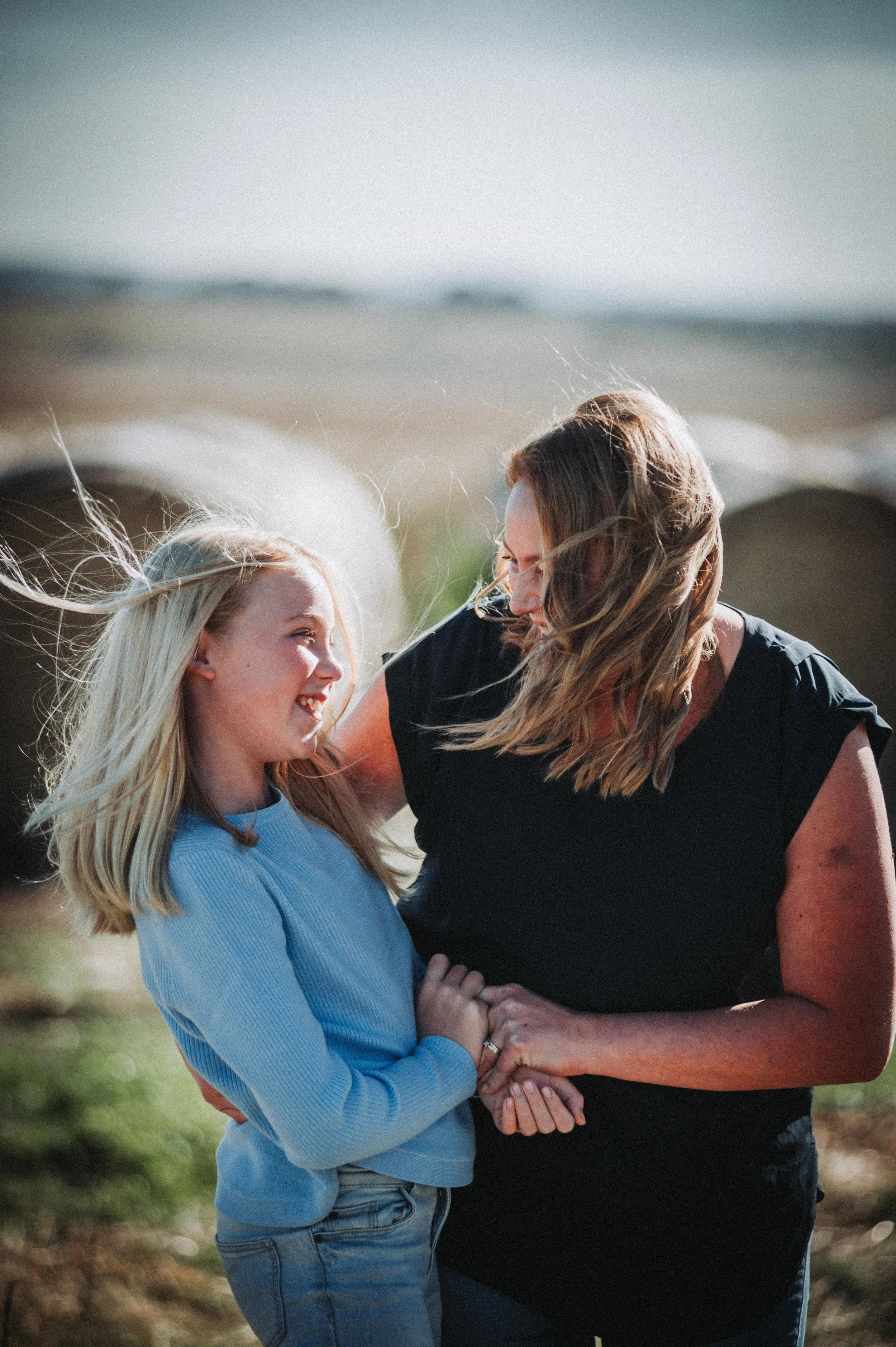 A woman and a young girl smiling at each other in a sunny outdoor setting with hay bales in the background.