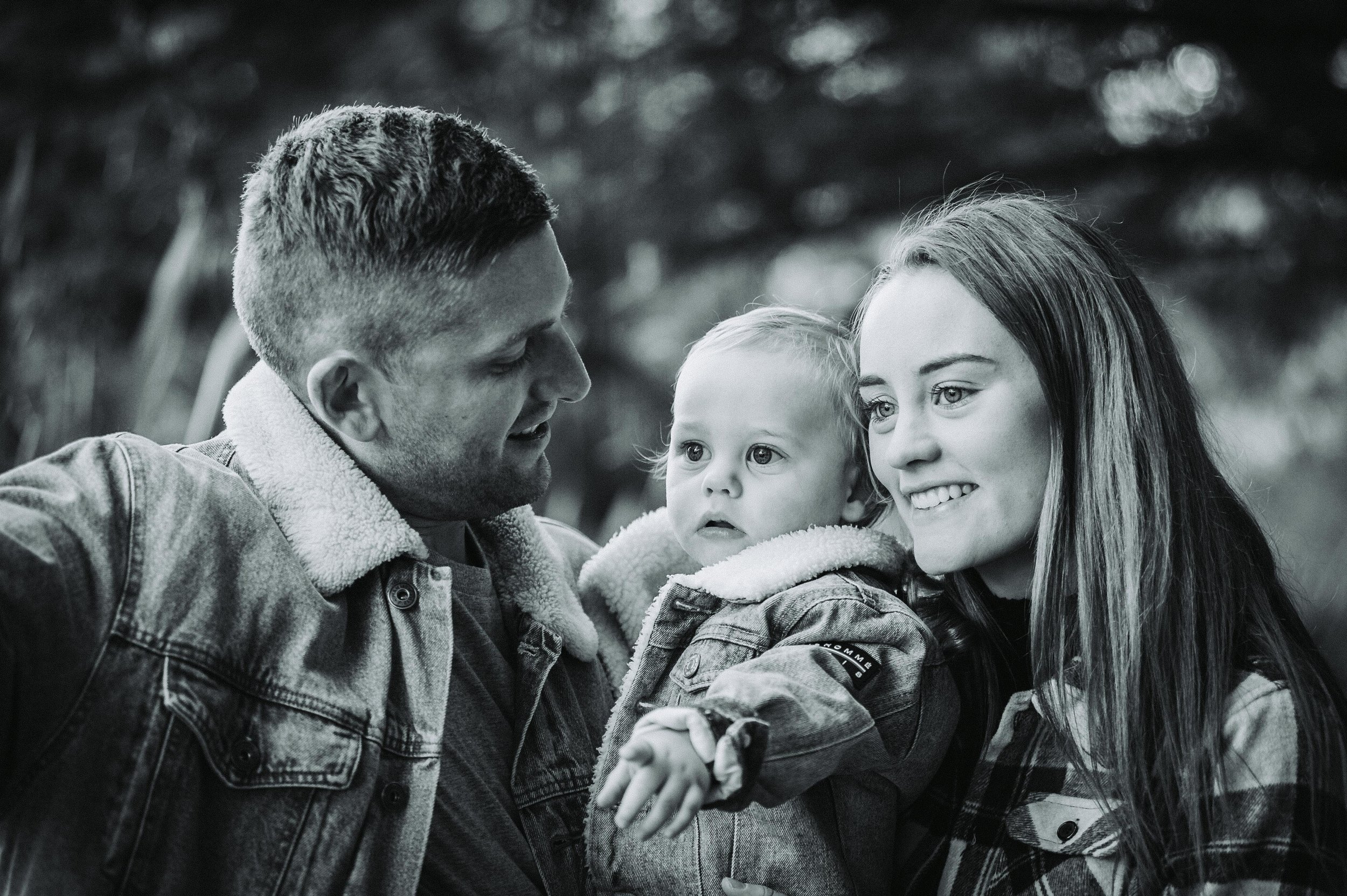 Black and white photo of a family with a man, woman, and child wearing denim jackets, smiling and looking in the same direction outdoors.