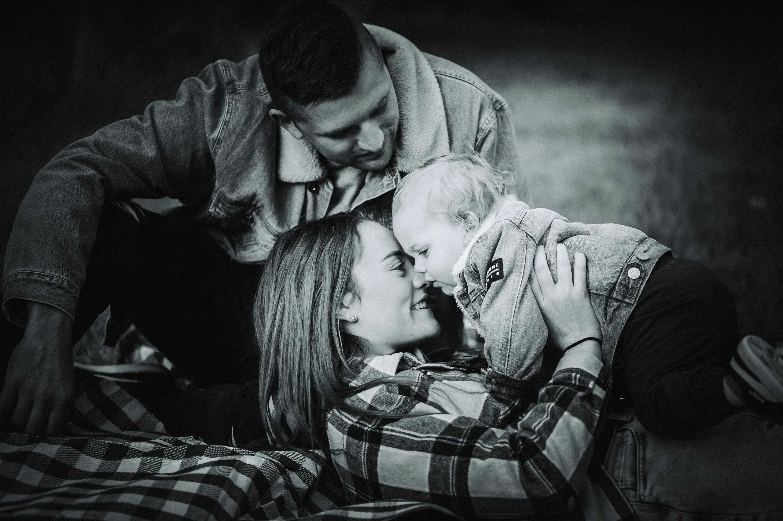 Black and white photo of a happy family outdoors, with a woman lying on a blanket, holding a baby, and a man leaning over them.