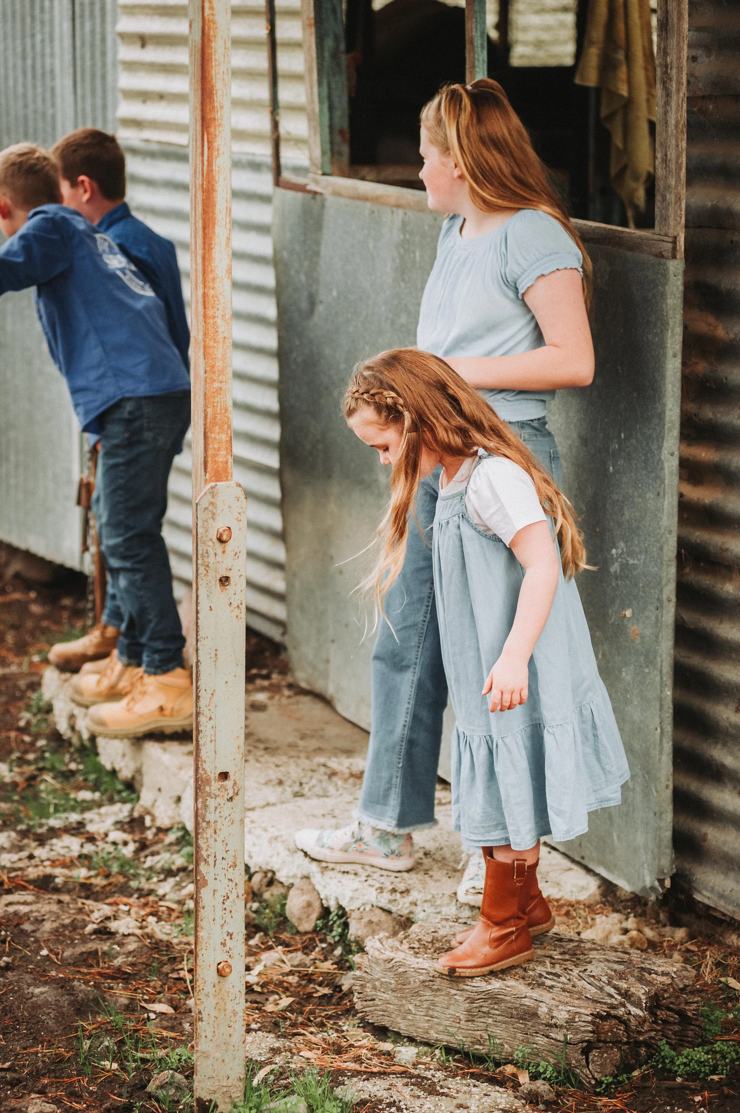 Three children standing outside a corrugated metal building, with two girls wearing blue outfits and a boy in a blue shirt and jeans. The girl in front is stepping down from a step, wearing brown boots.