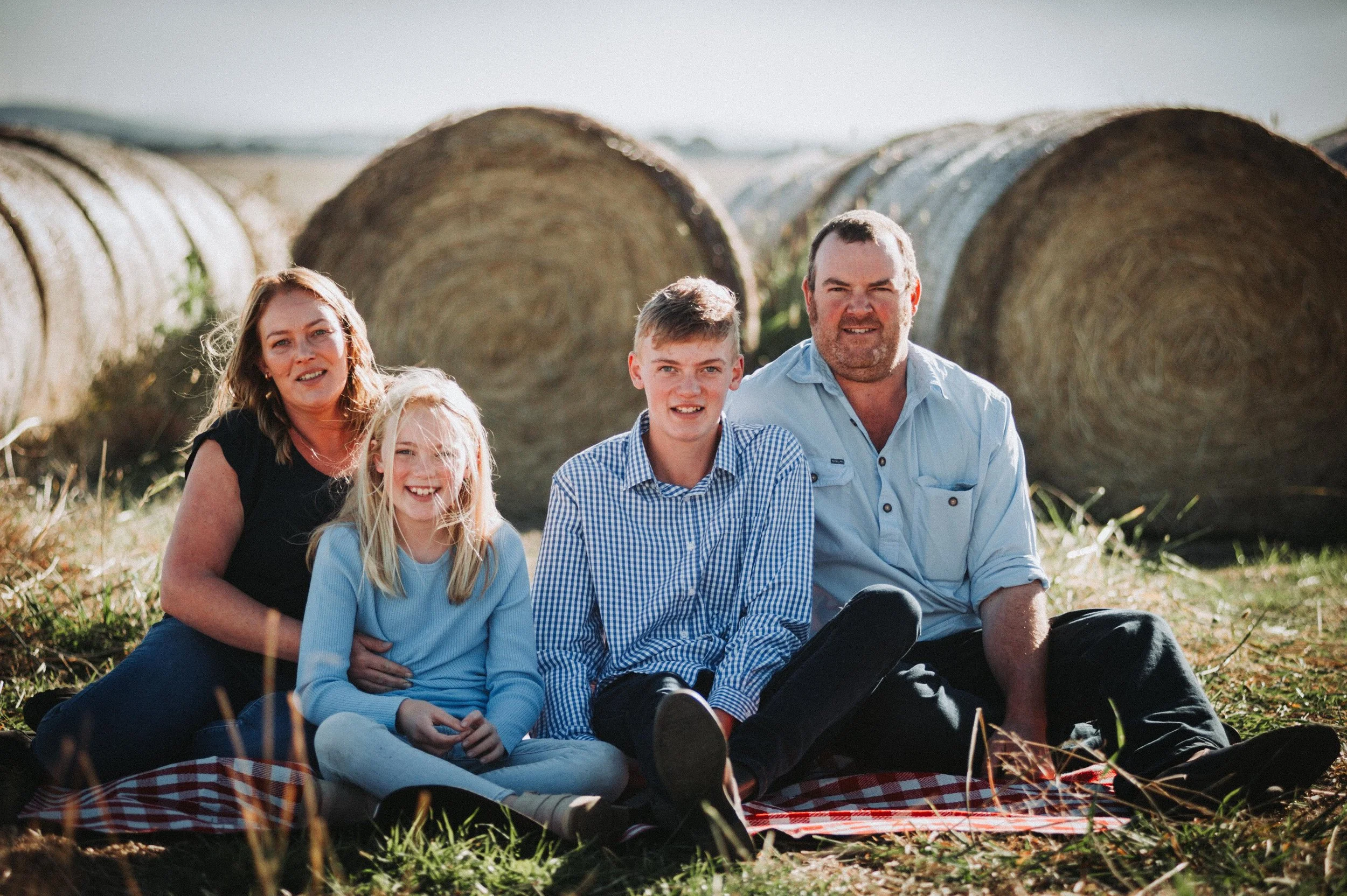 Family sitting on a picnic blanket in a field with hay bales in the background.