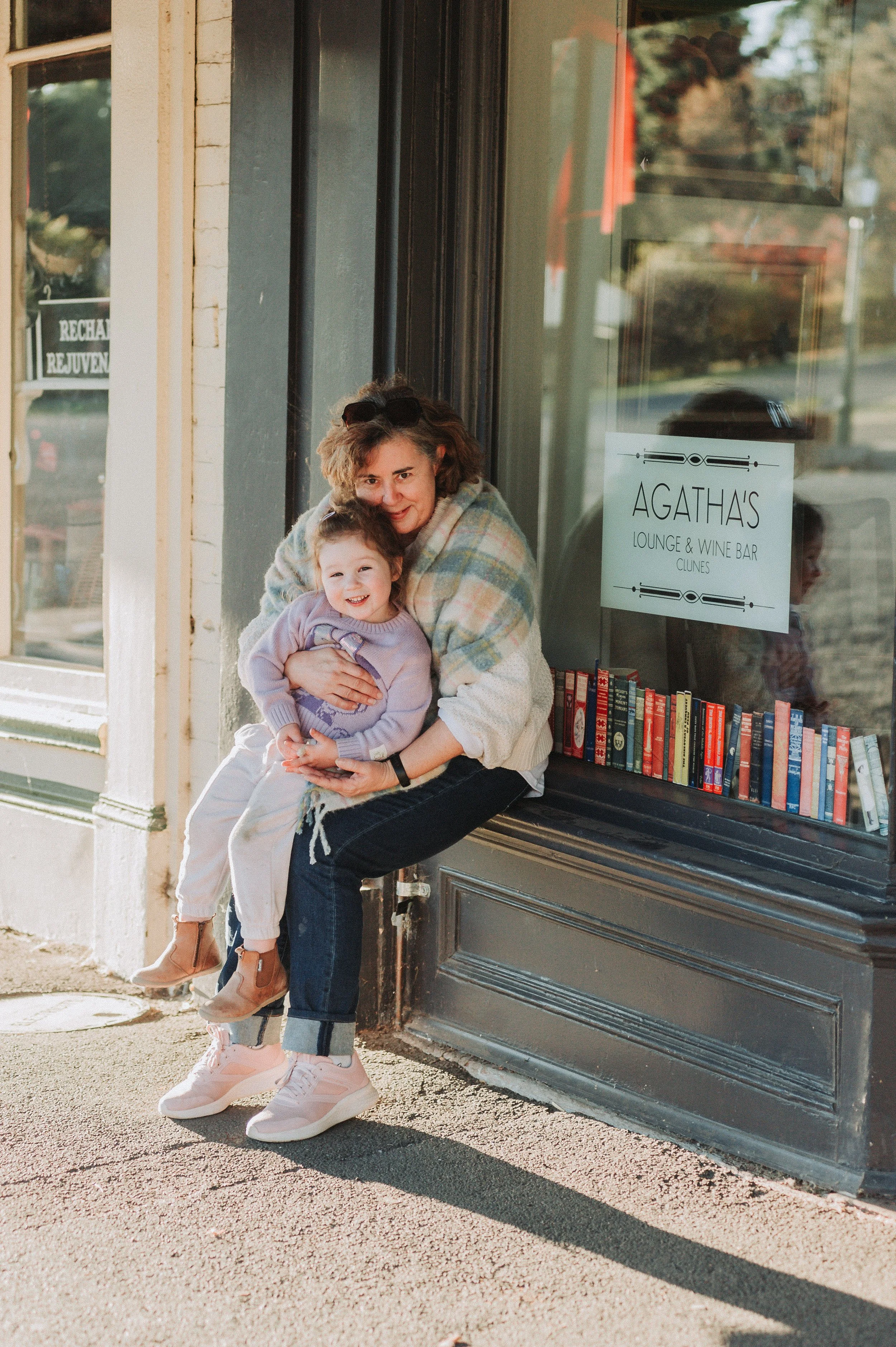 A woman and a young girl sitting outside a building with a sign that reads "Agatha's Lounge & Wine Bar Clunes." The woman is holding the girl on her lap, both smiling. Books are displayed on the windowsill behind them.