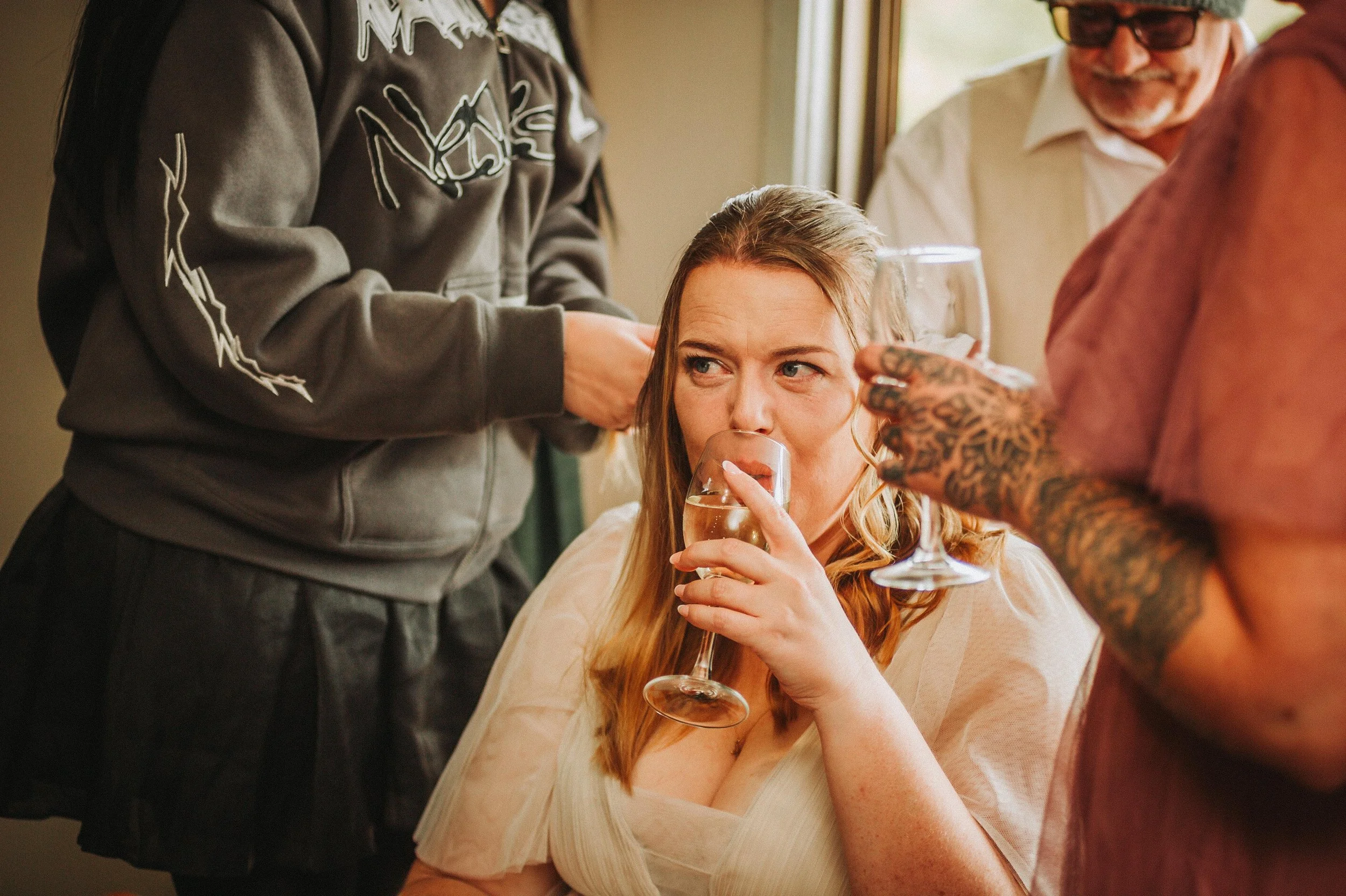 Woman in white dress drinking wine, surrounded by people, one styling her hair and another holding a wine glass.