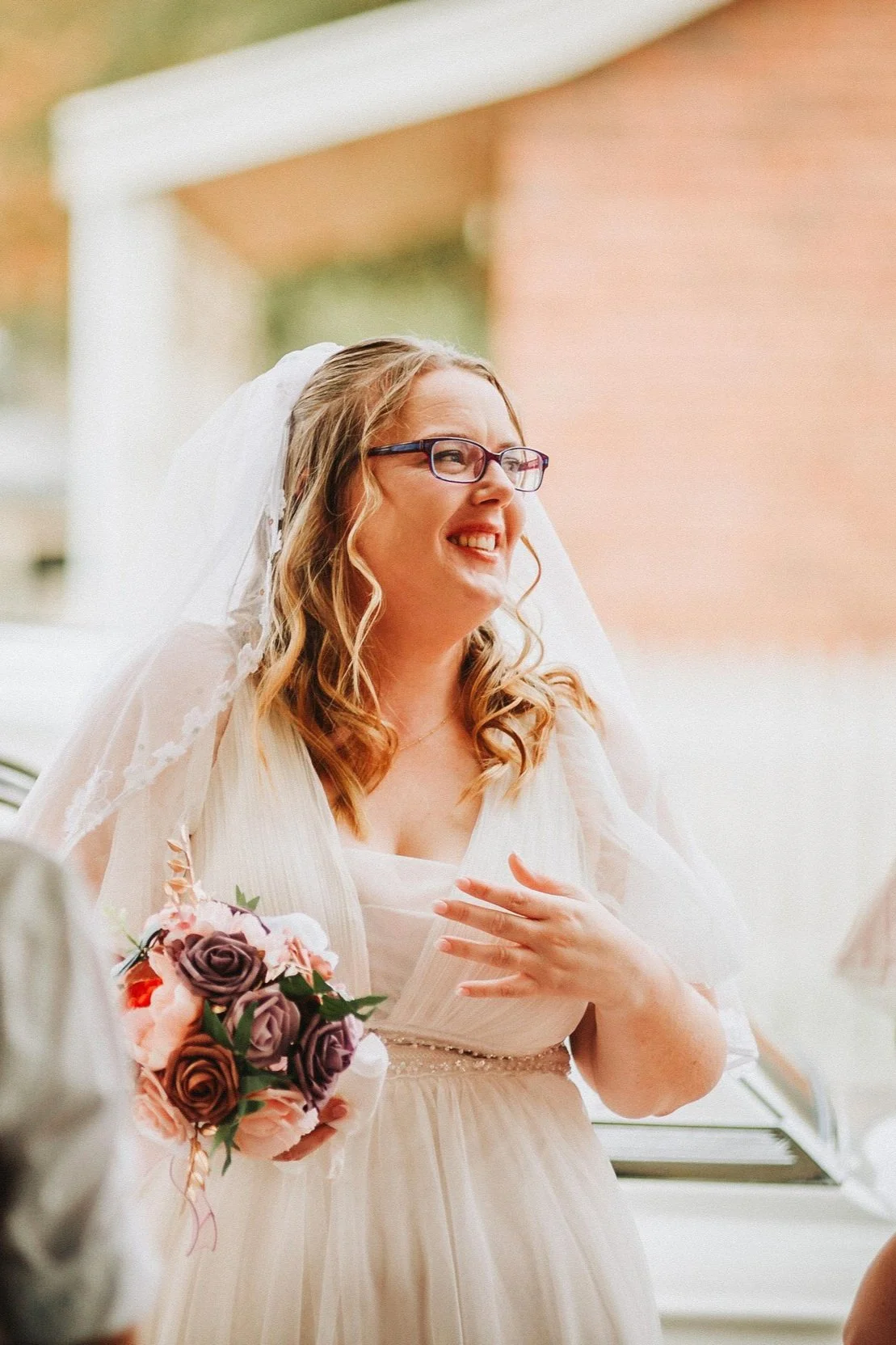 Bride smiling and holding a bouquet, wearing a veil and glasses