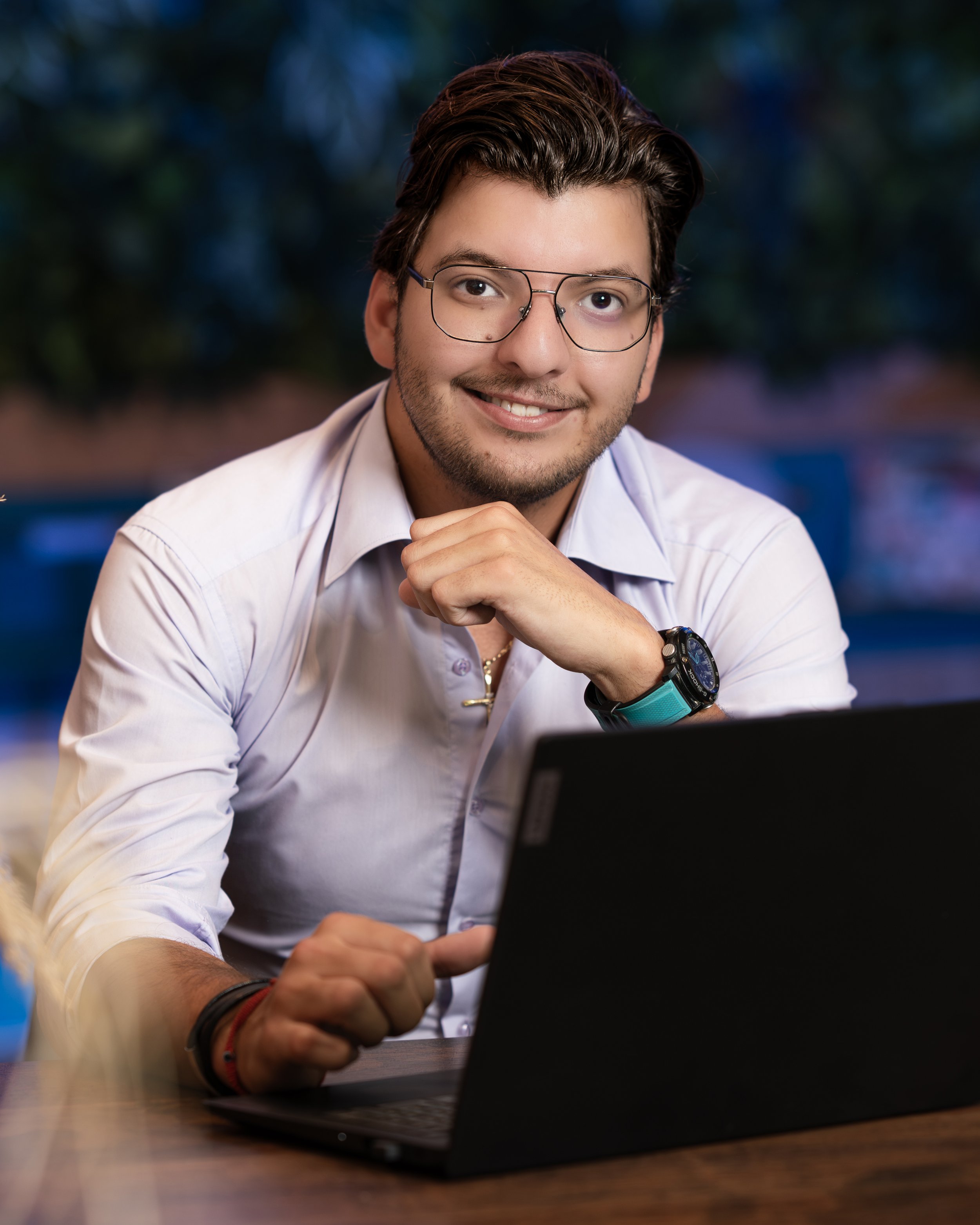A young man with glasses and dark hair, smiling, seated at a desk with a black laptop in front of him, outdoors at dusk.