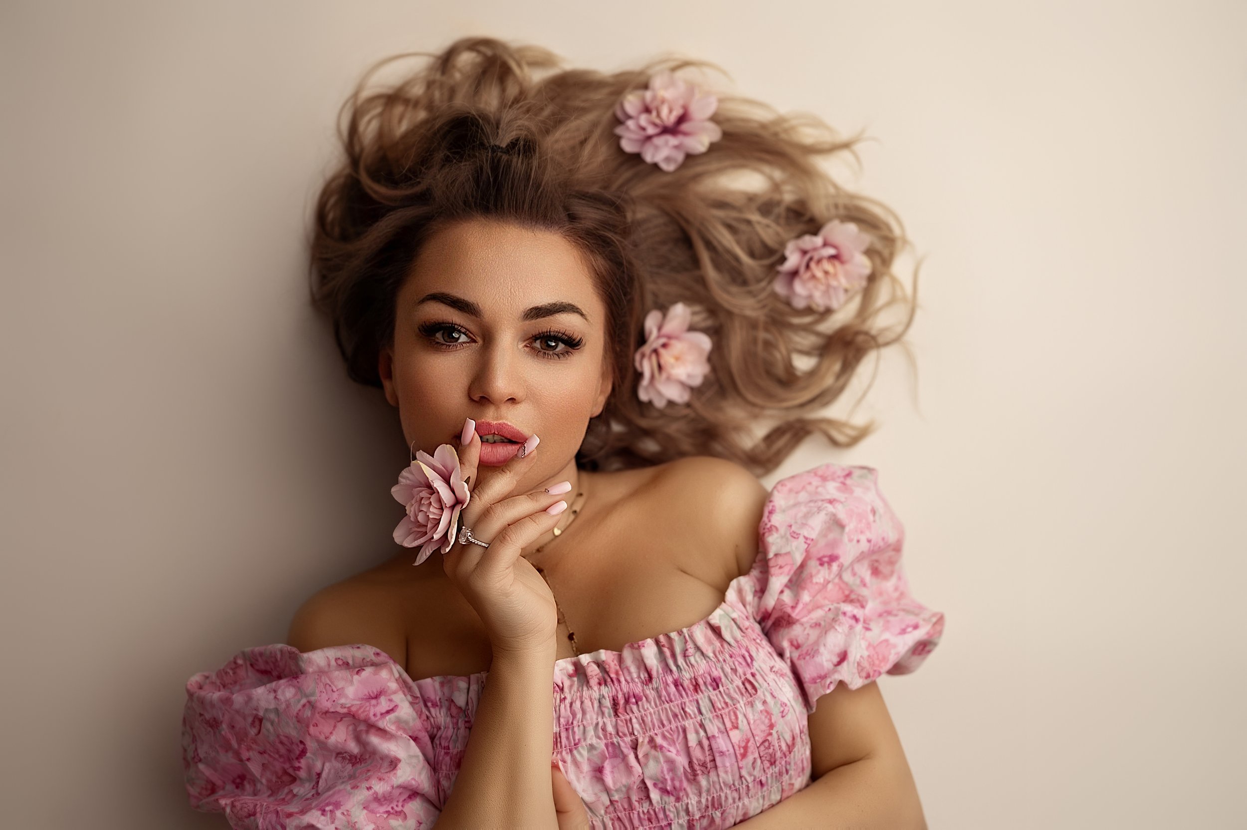 A woman with wavy blonde hair adorned with pink flowers, wearing a pink floral off-shoulder dress, gently touching her lips with her left hand.