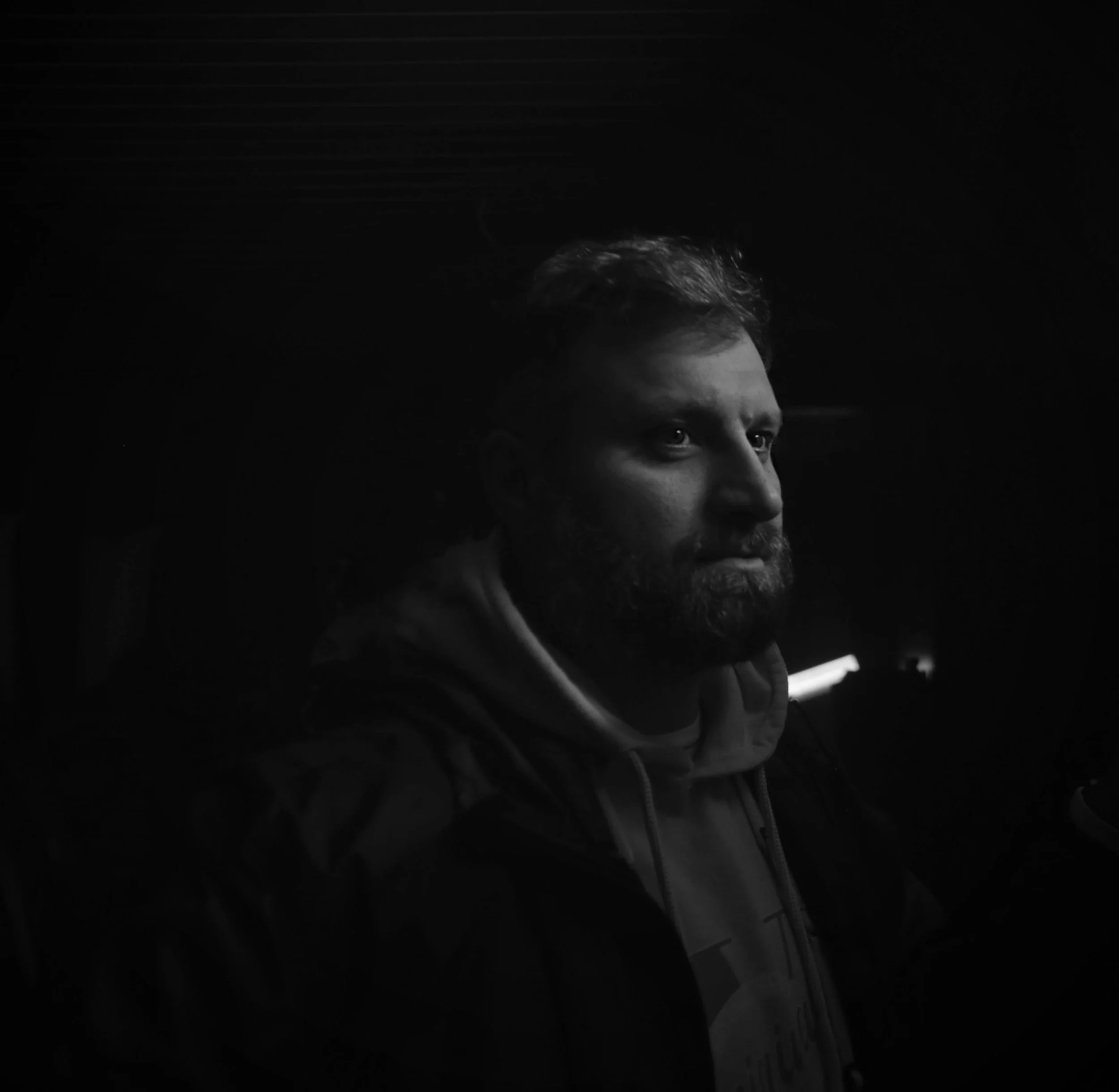 Black and white photo of a serious man with a beard, looking to the right, in a dark environment with minimal lighting.