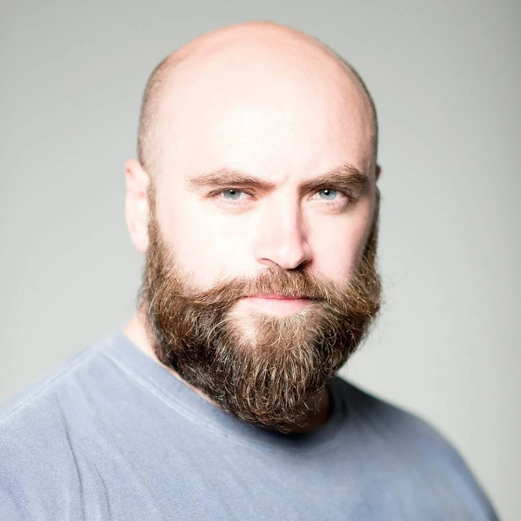 A close-up portrait of a bearded man with light skin, blue eyes, and a serious expression, wearing a gray shirt against a plain background.