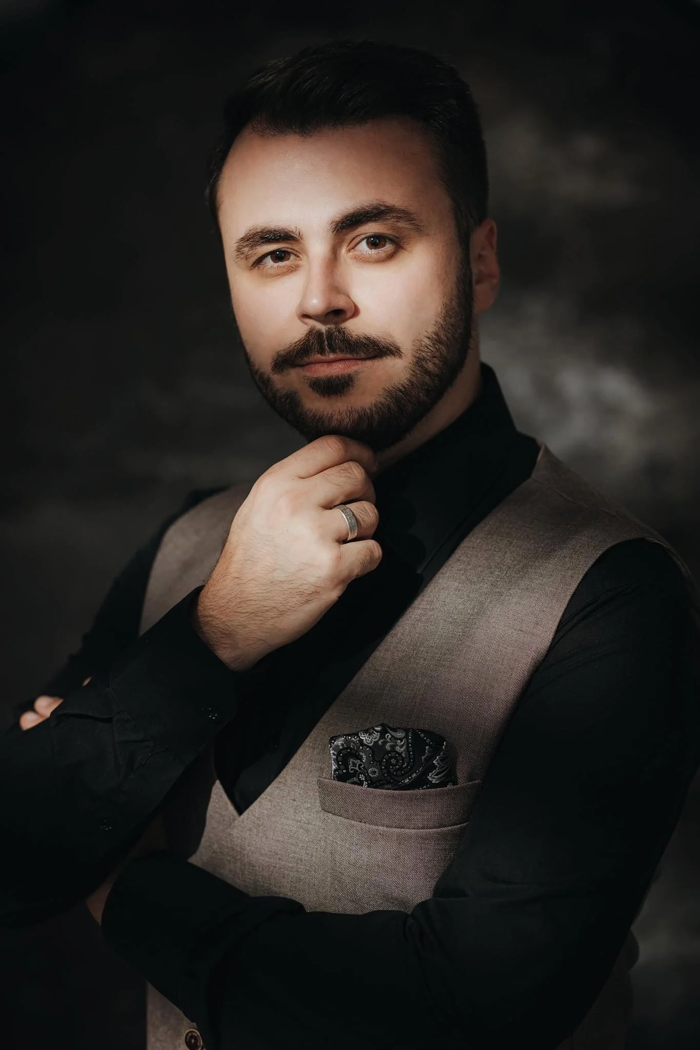 A man with dark hair and a beard, wearing a black shirt and a gray vest, with a pocket square, posing with his hand on his chin, against a dark, blurred background.
