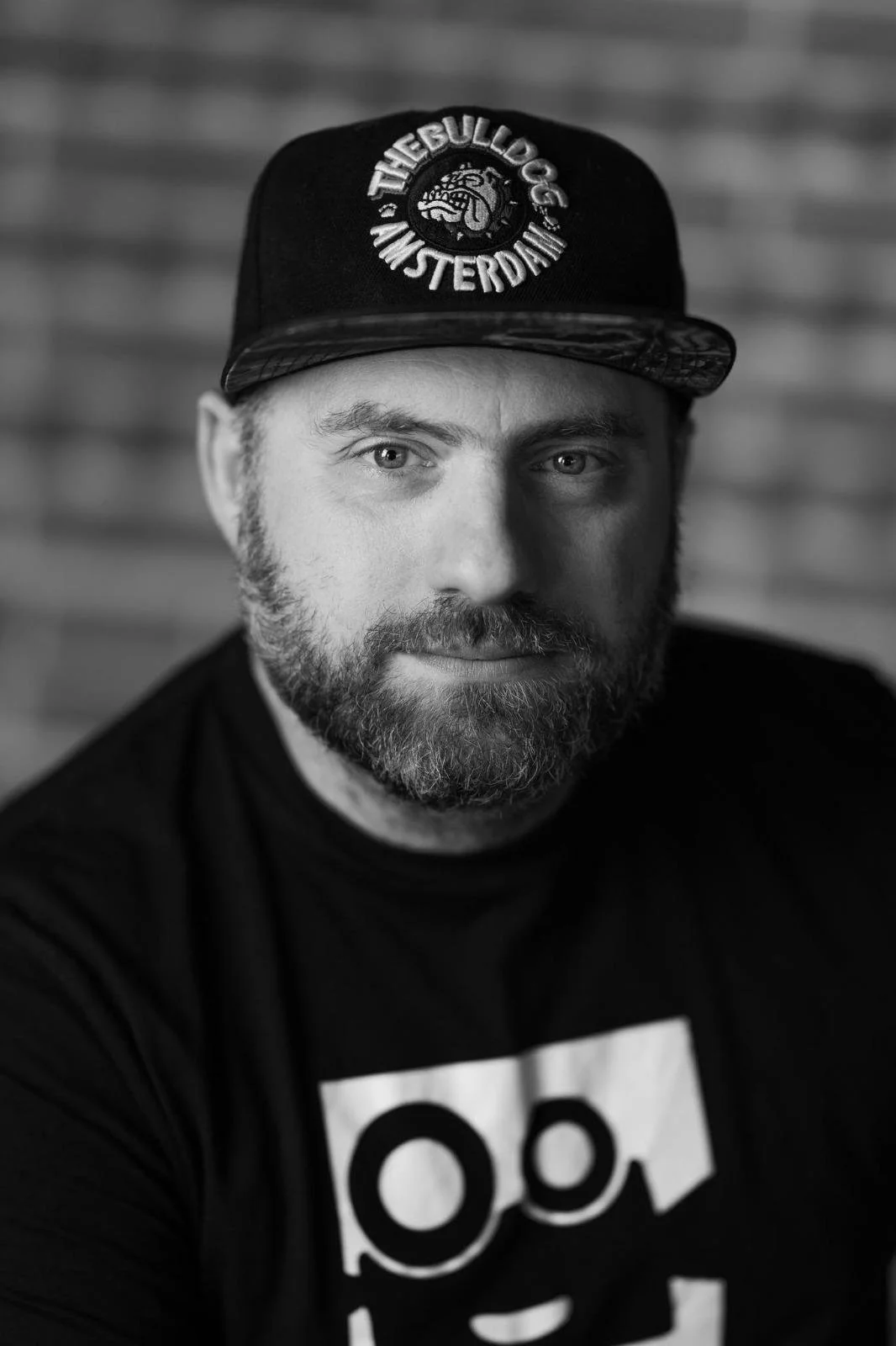Black-and-white portrait of man with a beard, wearing a black cap with logo and text 'THEBULLDOG AMSTERDAM' and a black T-shirt with a graphic design.