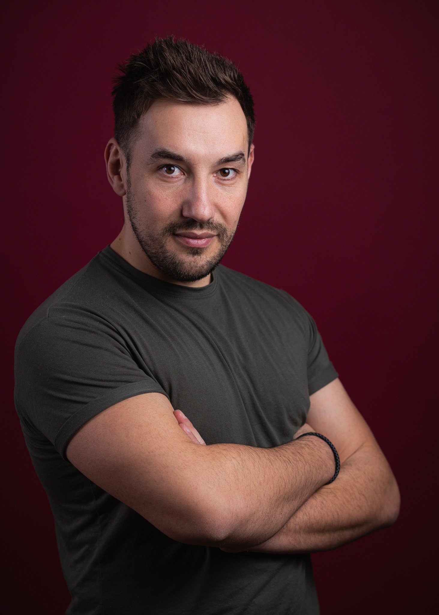 A man with dark hair and a beard, wearing a dark t-shirt, standing with arms crossed against a maroon background, looking at the camera.