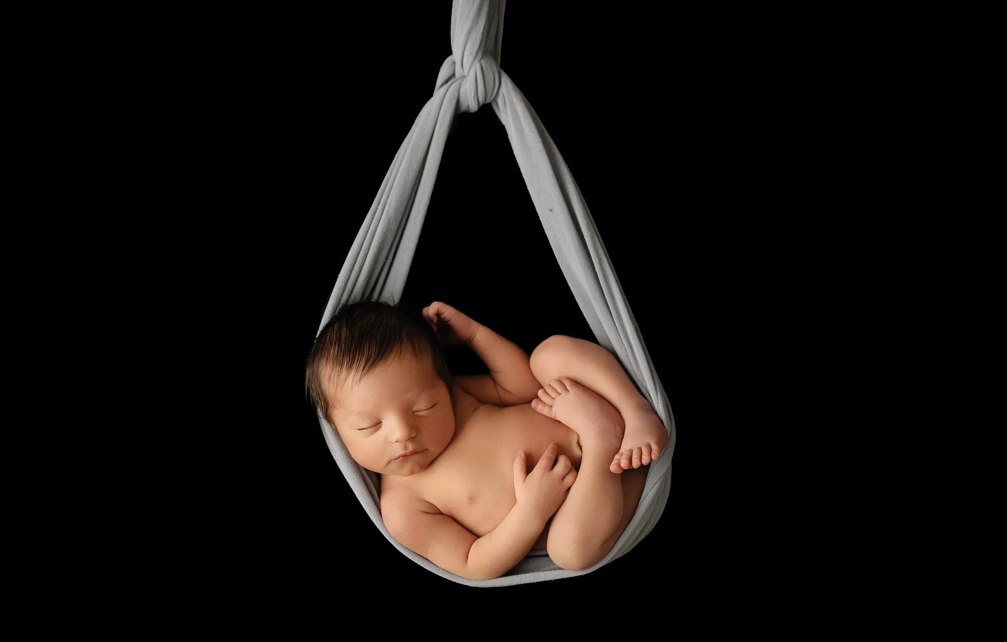 A newborn baby peacefully sleeping in a gray fabric hammock against a black background.