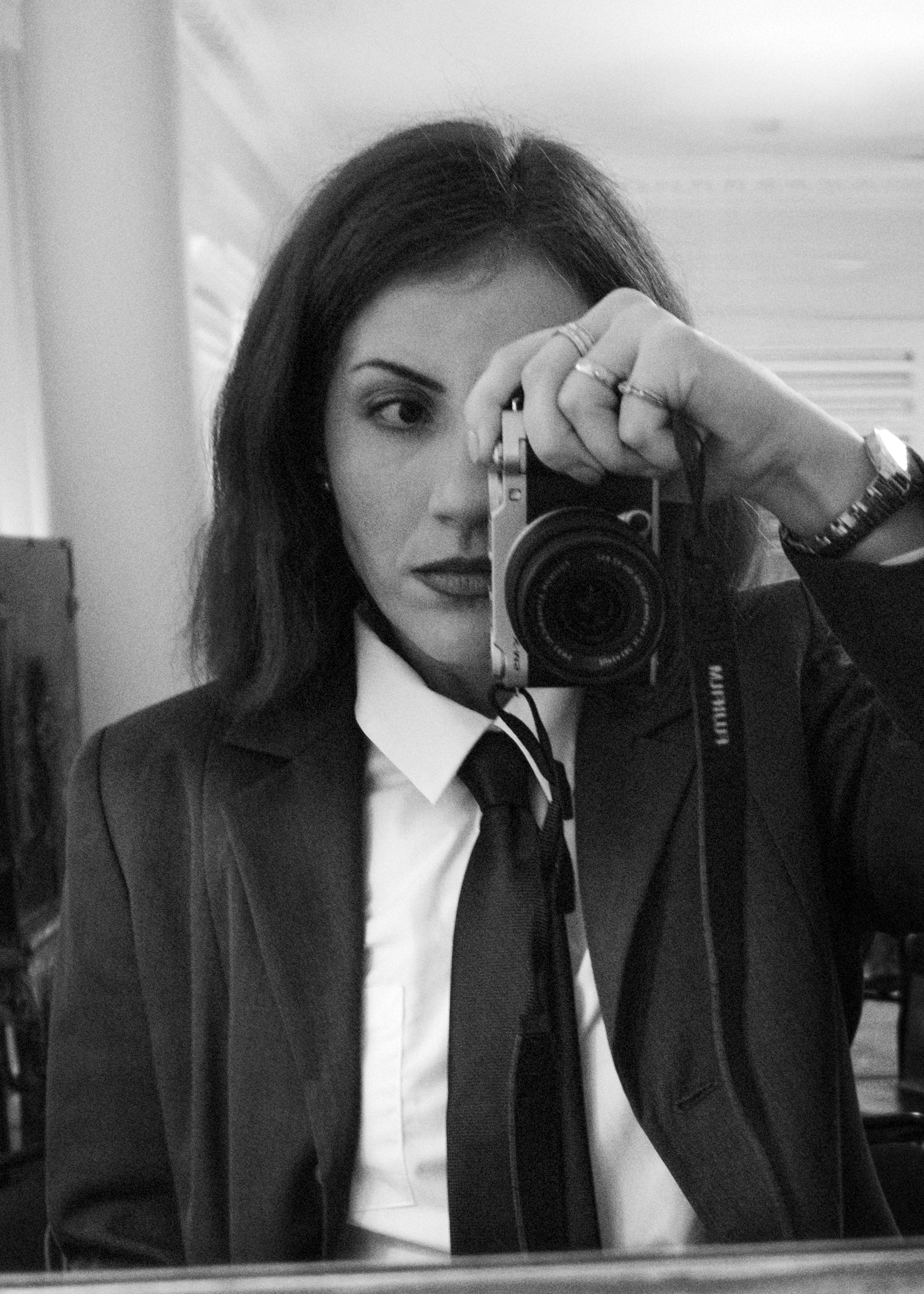 A woman with dark hair, wearing a suit and tie, takes a black-and-white selfie in a mirror with a camera, partially covering her face.