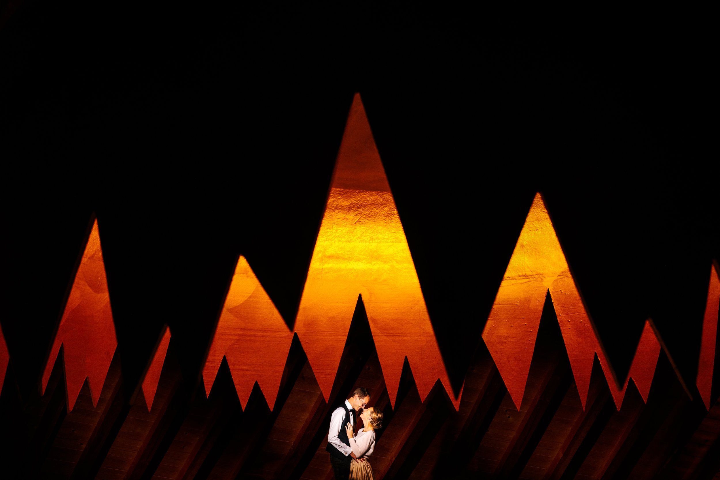 A couple standing close together and looking into each other's eyes beneath orange-lit, triangular-shaped decorations hanging from the ceiling.