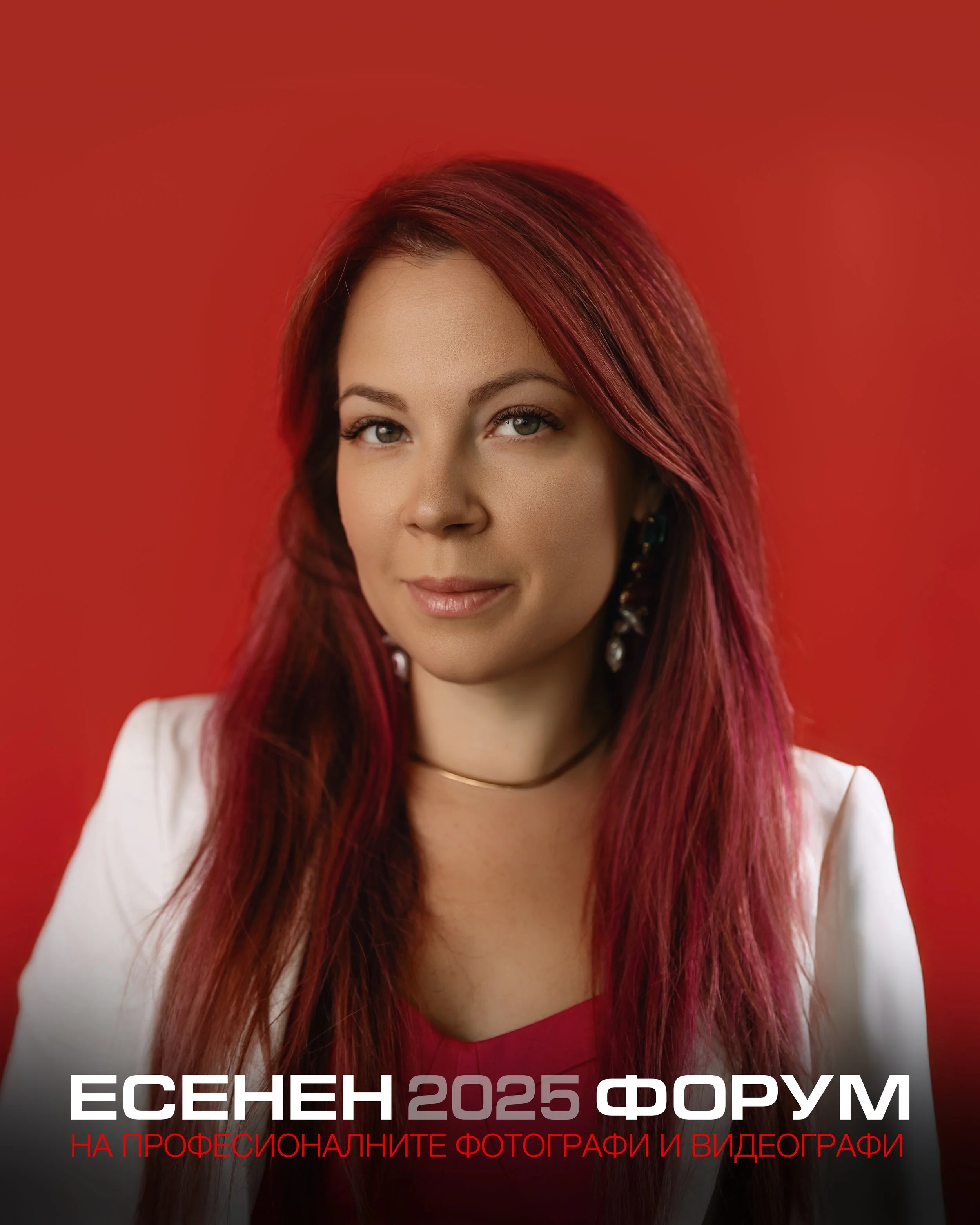 A woman with red hair and light skin smiling against a red background, wearing earrings, a white blazer, and a red top.
