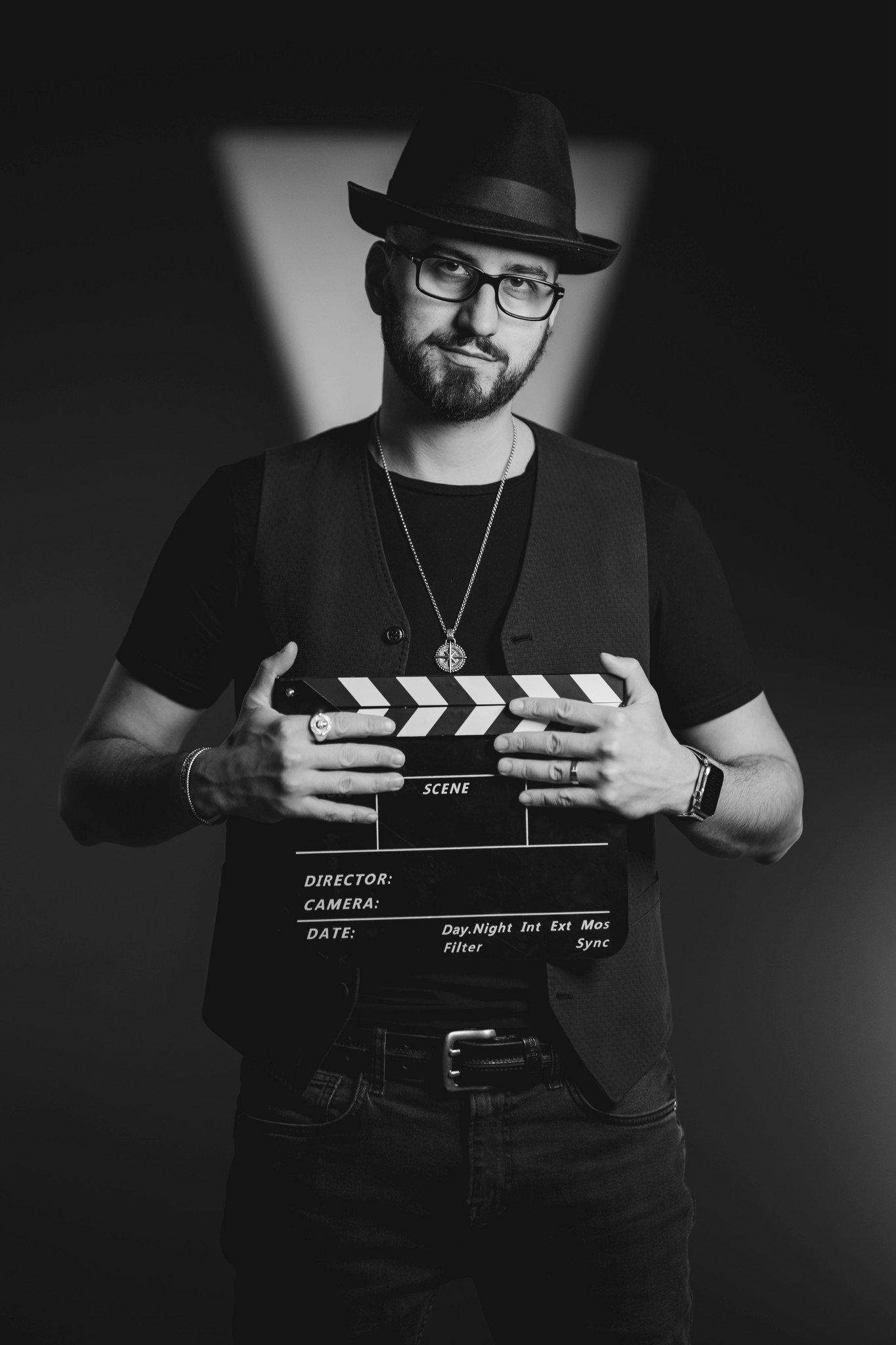 A man wearing a hat, glasses, and a necklace, holding a film clapperboard in a studio with dramatic lighting.