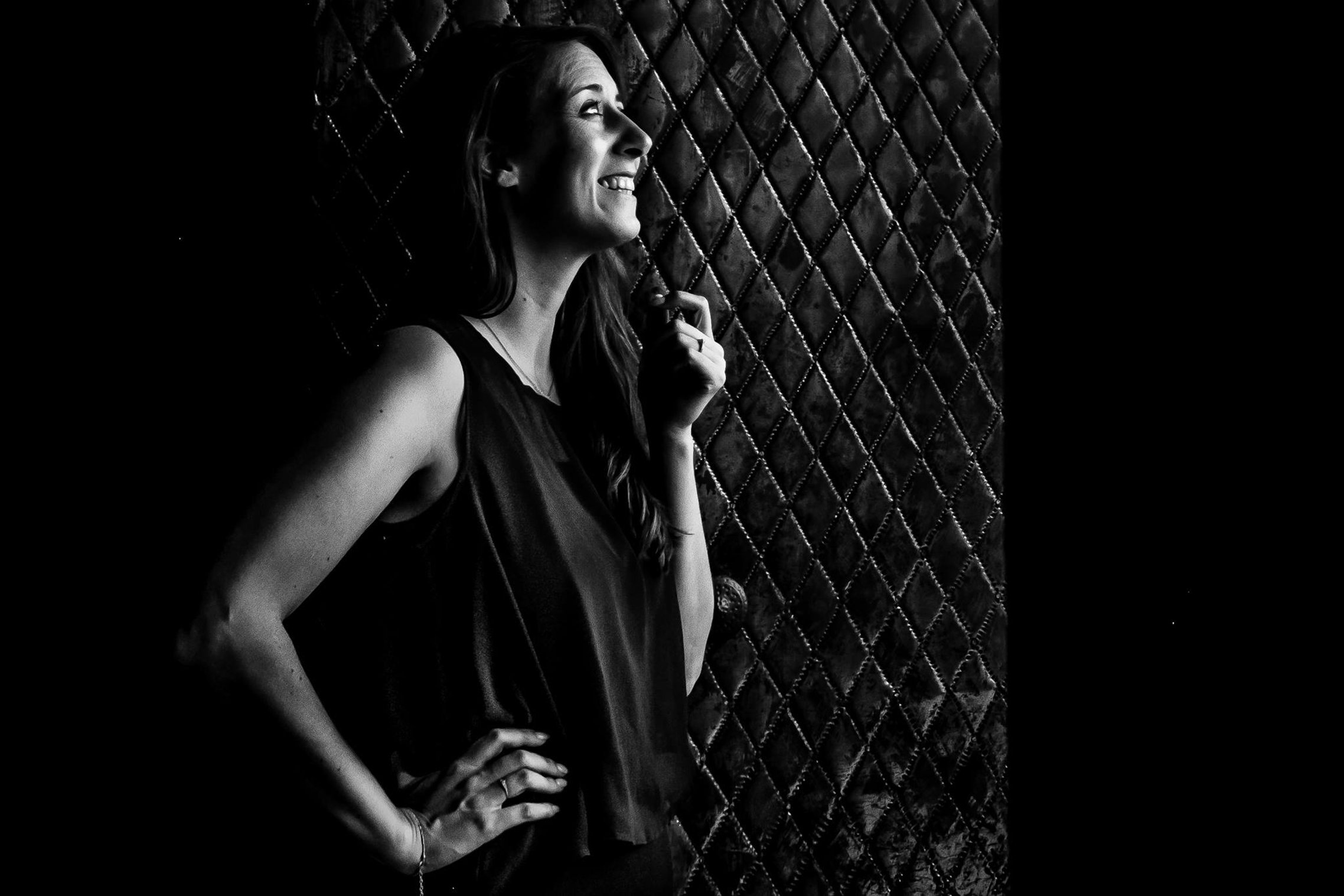 A black and white photo of a woman with long hair, smiling and looking to her left, standing with one hand on her hip and the other lightly touching her shoulder, in front of a quilted background.