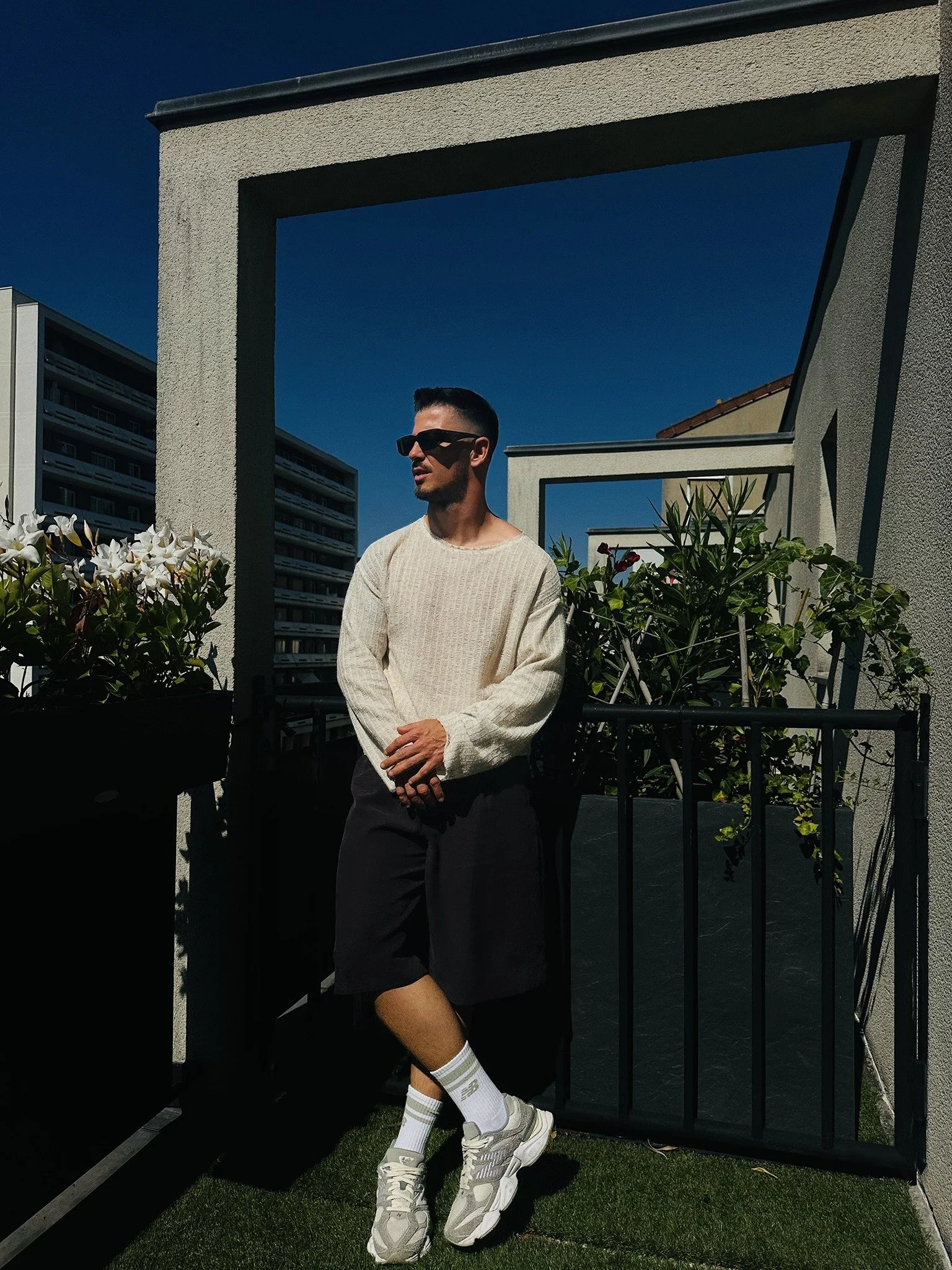 A man wearing sunglasses, a white sweater, black shorts, white socks, and sneakers standing on a small balcony with plants, framed by architectural rectangular structures, against a clear blue sky.
