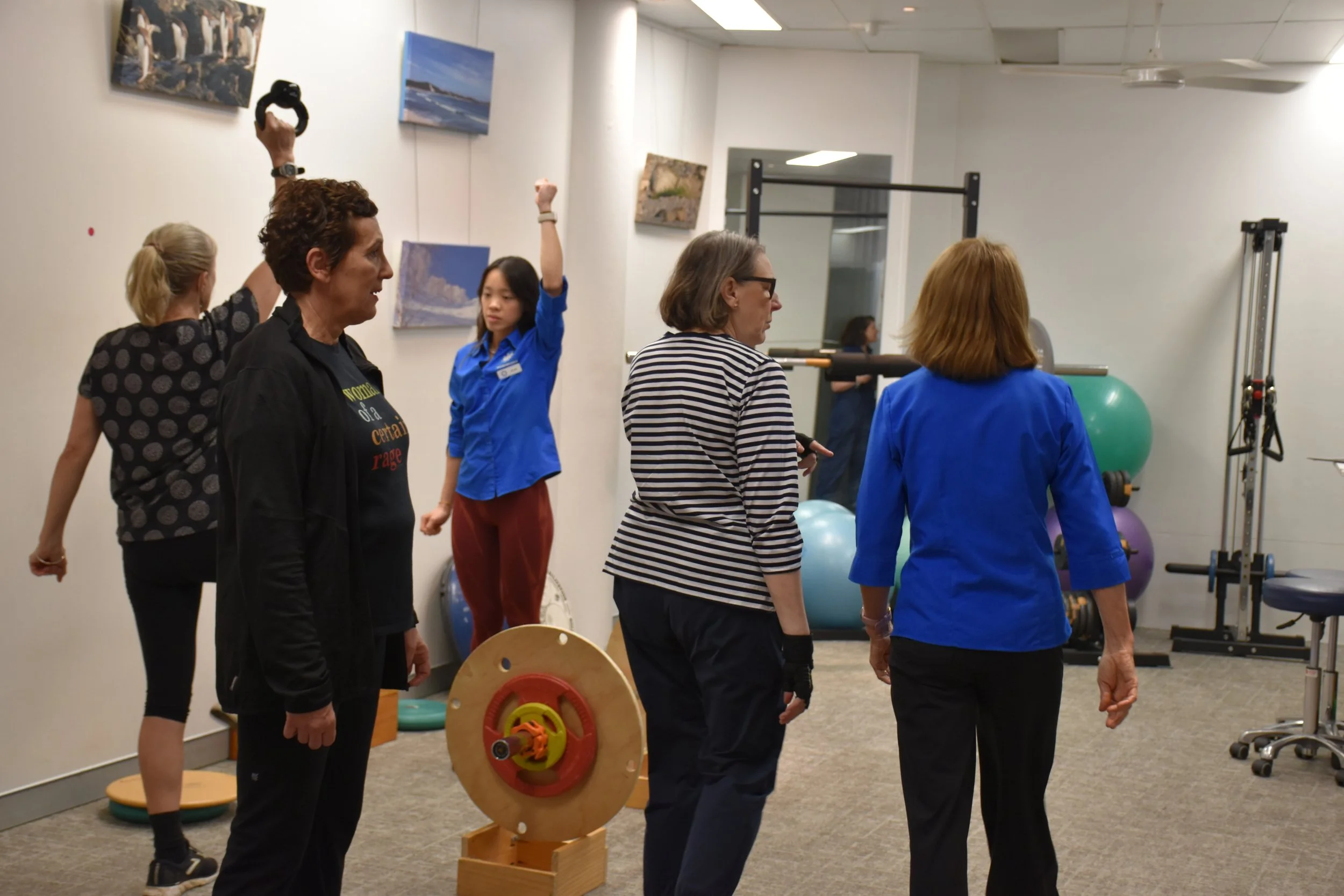 A physiotherapist leads a participant somewhere while another physiotherapist shows another clint how to hold a weight over her head in the background