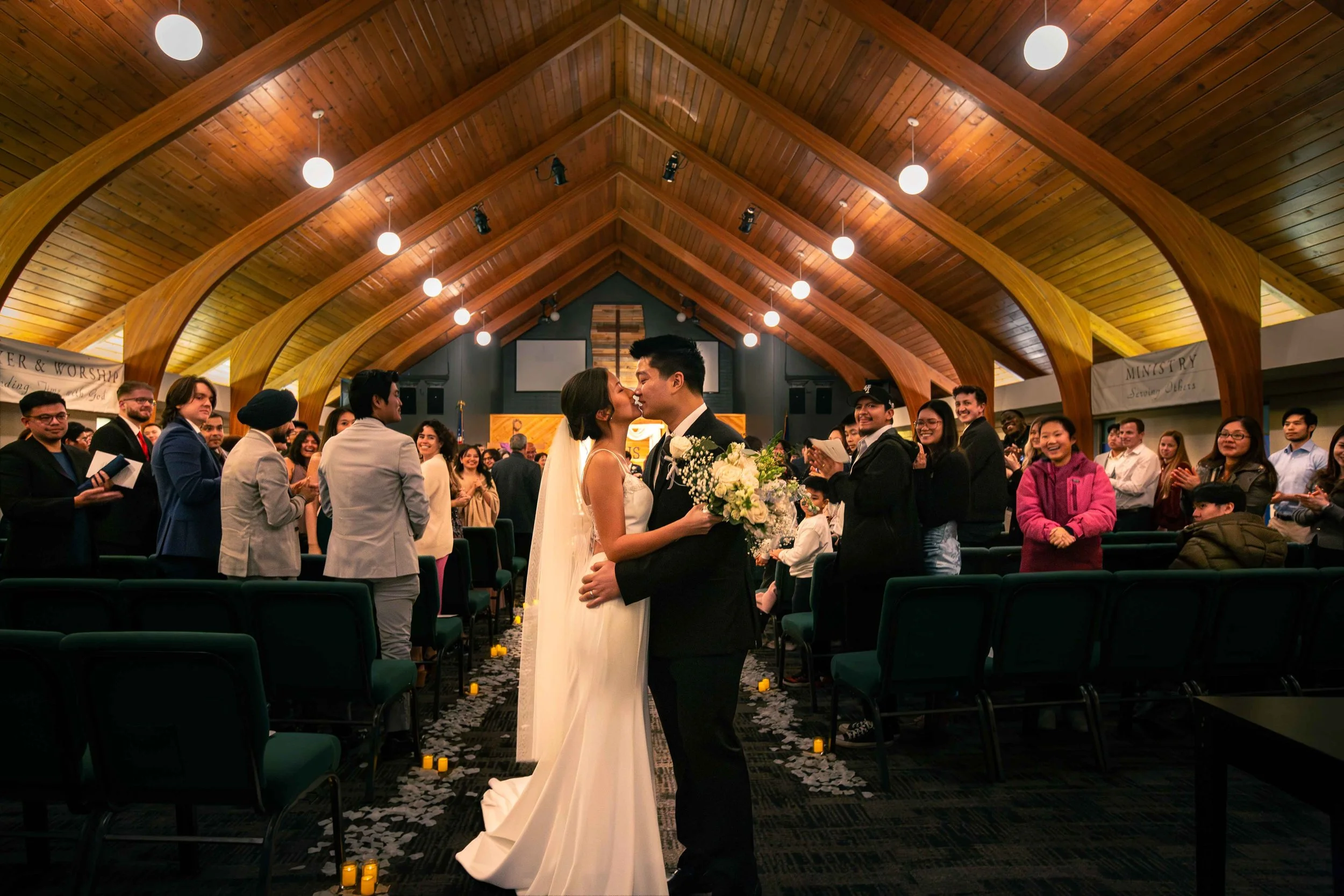 A bride and groom kiss at their wedding ceremony inside a church or chapel with a wooden vaulted ceiling. Guests stand on either side, clapping and smiling, with some holding programs. The aisle is decorated with small candles and flower petals.