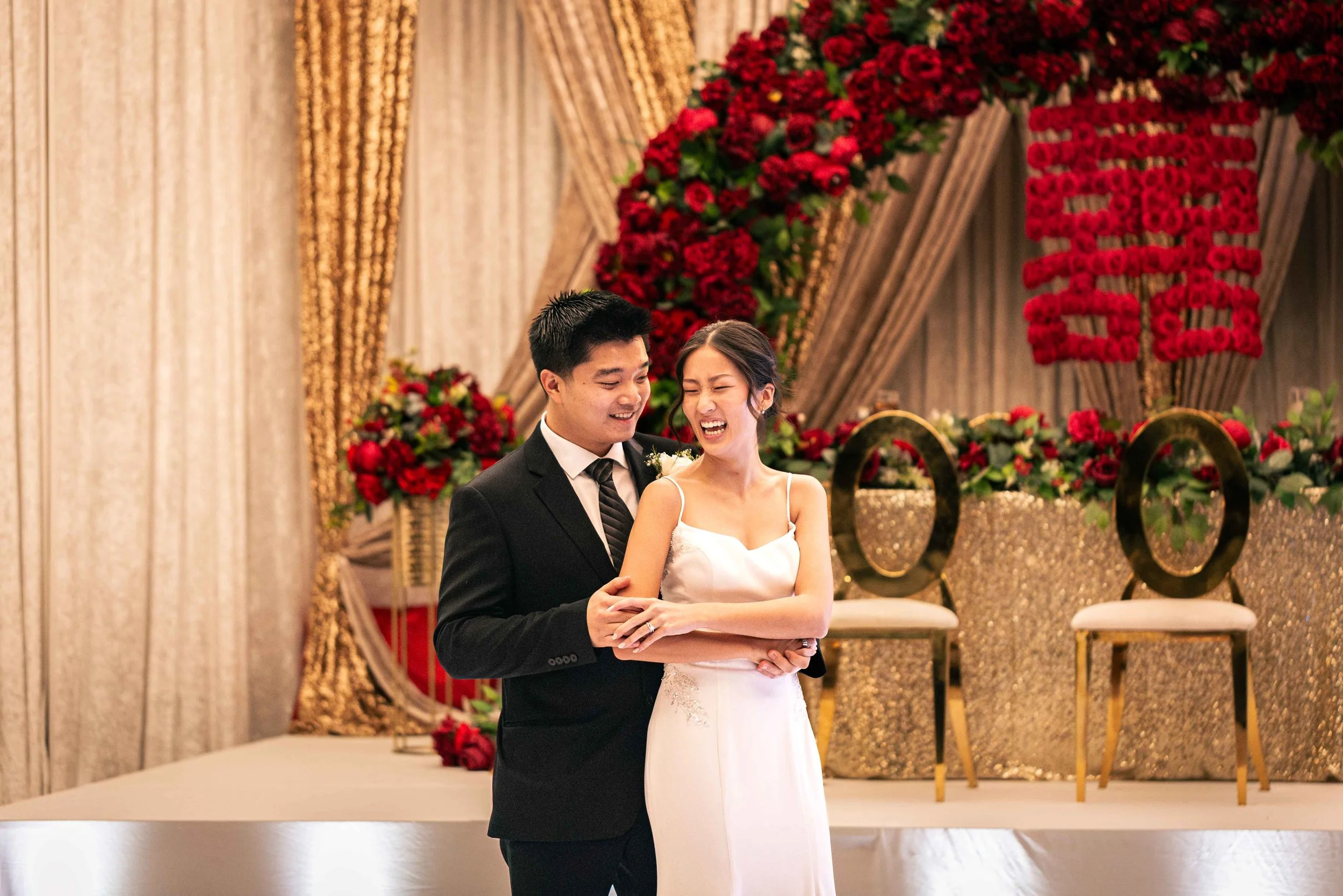 A smiling bride and groom dance together at their wedding reception, with a floral backdrop featuring red roses and gold accents.