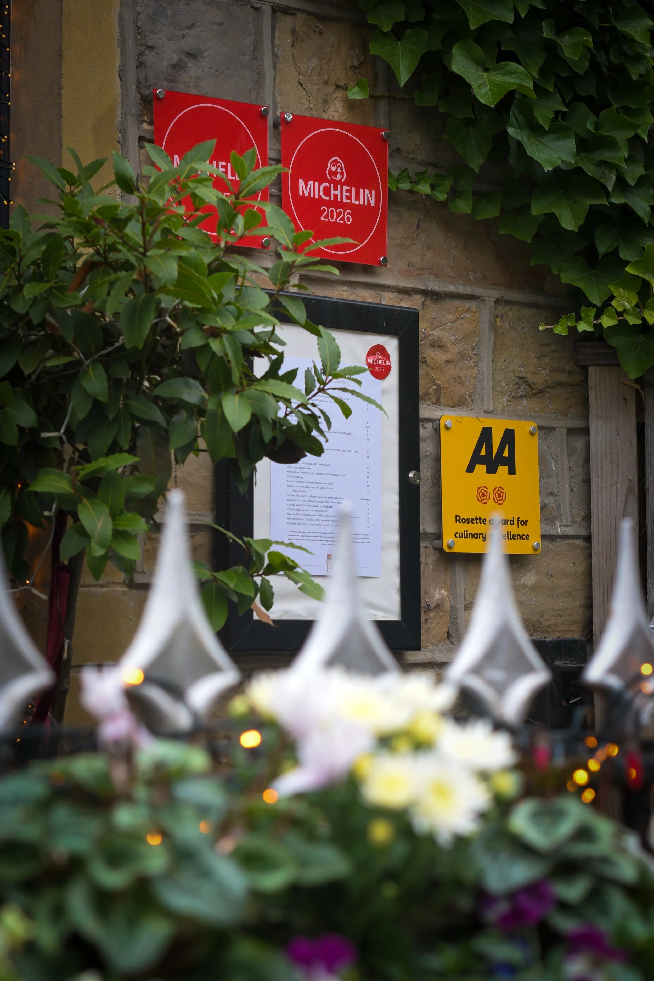 Outdoor scene featuring green ivy and other plants on a stone wall. Several signs are mounted on the wall, including red Michelin 2026 award plaques, a framed menu, and a yellow AA plaque. The foreground is blurry with flowers and decorative lights.
