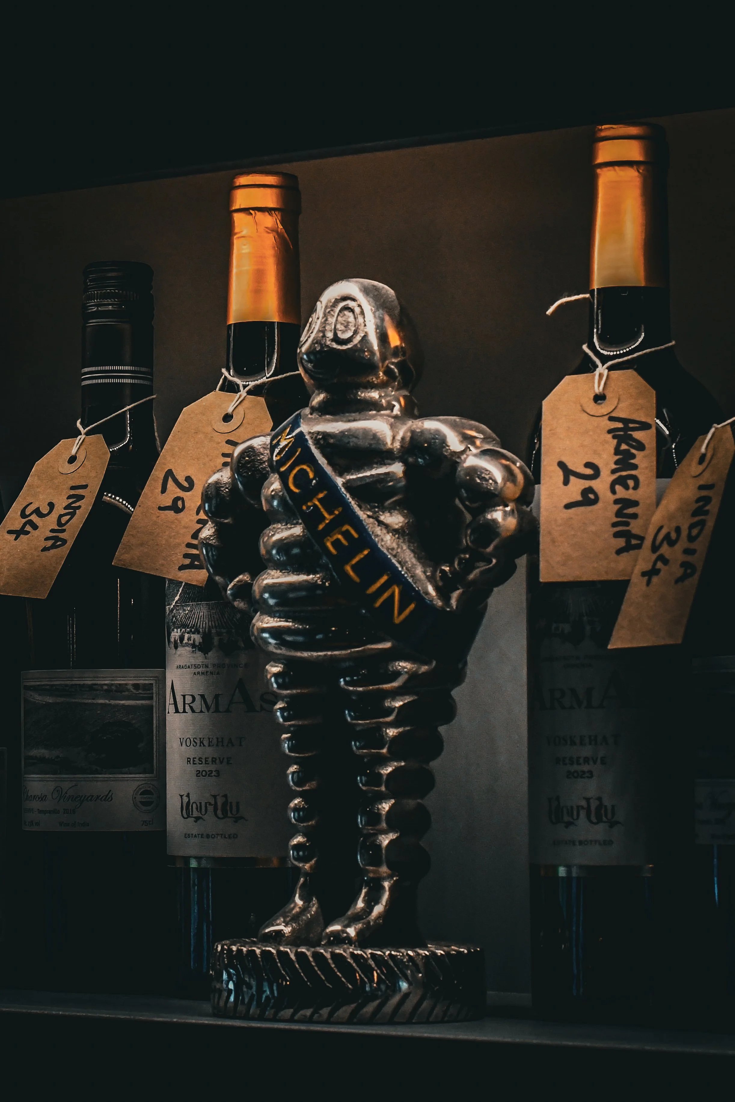 A metal sculpture of a vintage motorcycle engine with a small figure of a person sitting on top, wearing a sash labeled 'MUCHELIN,' displayed in a glass case alongside bottles of wine with tags.