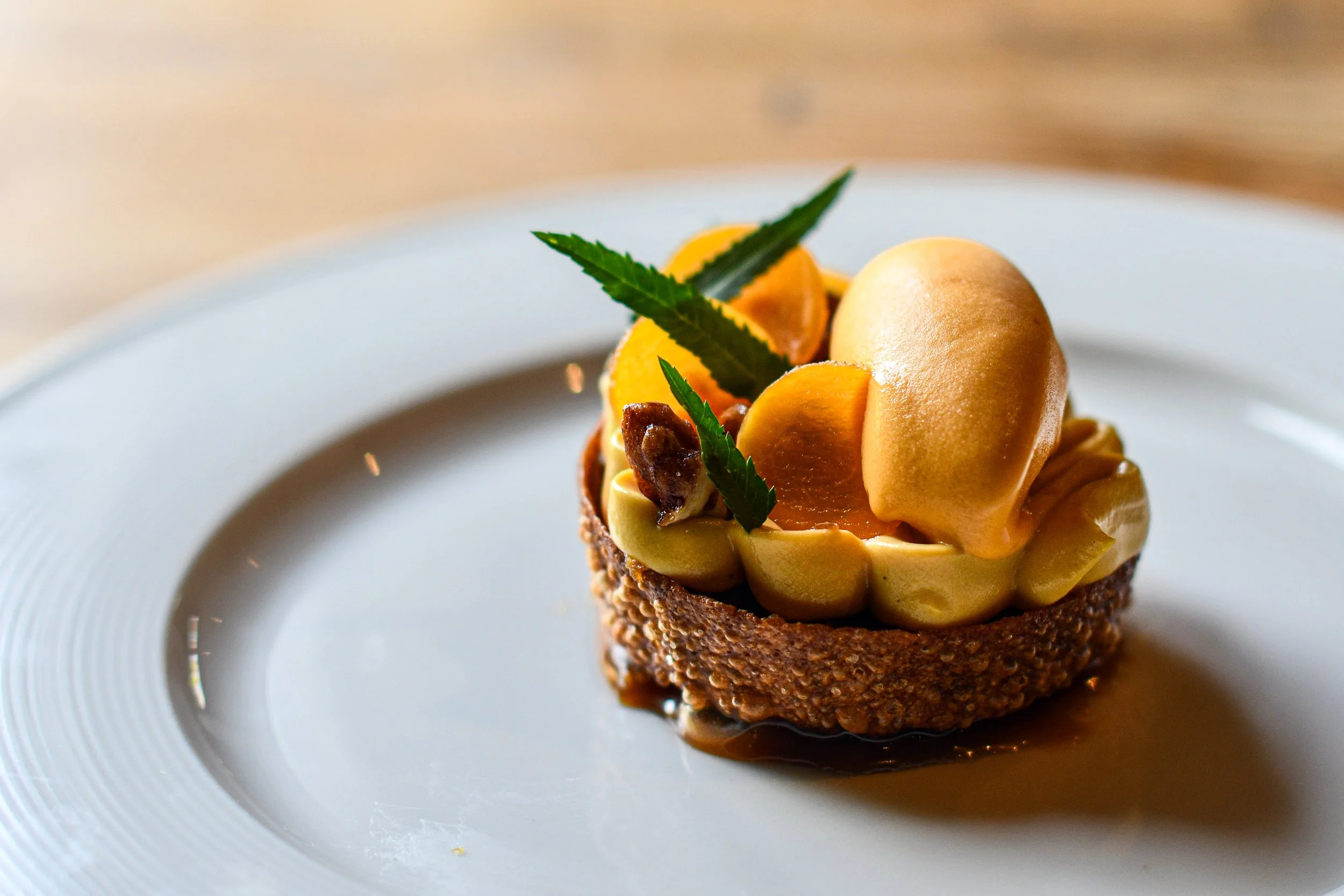 A plated dessert featuring a tart crust filled with creamy filling, topped with slices of fruit, garnished with green leaves, and drizzled with caramel sauce.