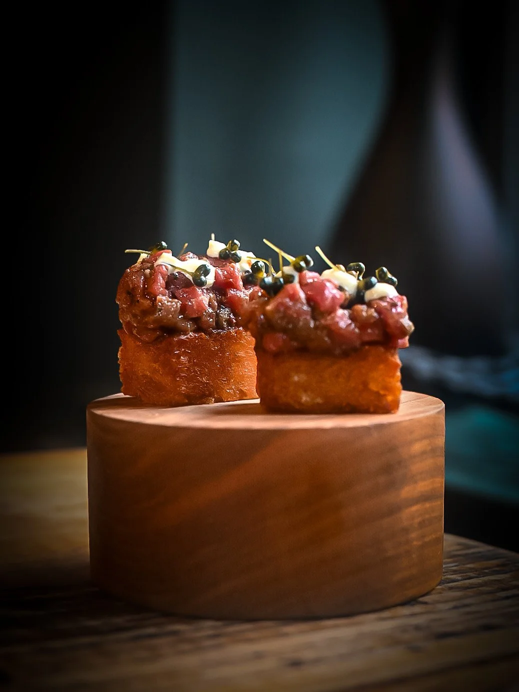 Two gourmet croquettes topped with a mixture of chopped ingredients and microgreens, served on a wooden block.