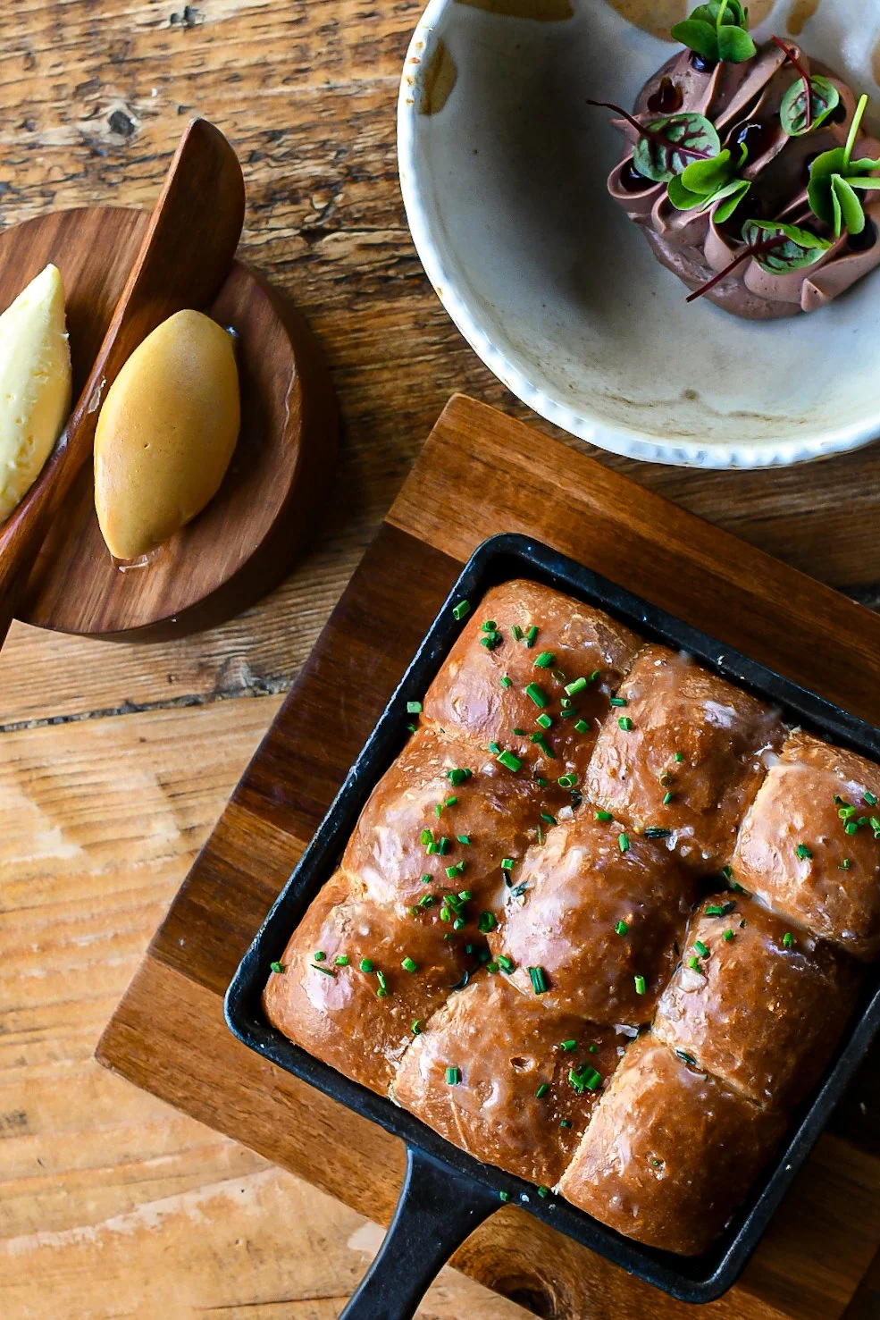 Cooked meatloaf garnished with chopped chives served in a baking dish, with a bowl of chocolate mousse topped with microgreens, and a wooden tray holding two scoops of mango ice cream.