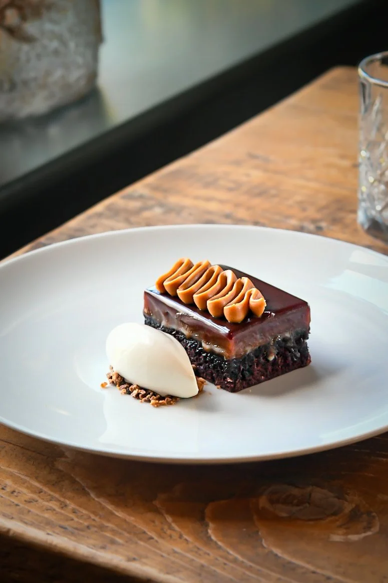 Chocolate layered dessert topped with caramel swirls, served with a dollop of whipped cream or mascarpone and garnished with chocolate shavings on a white plate.