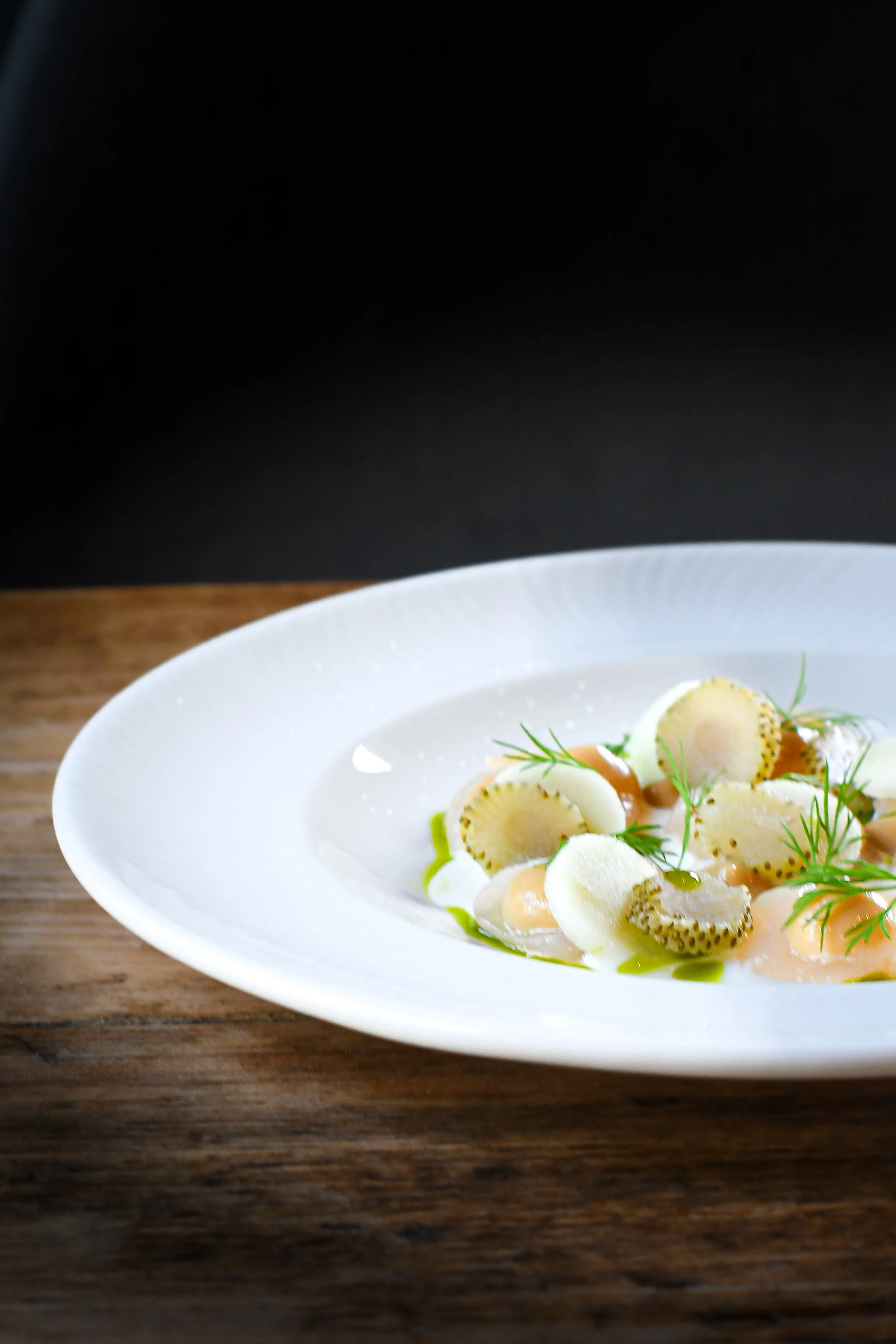 Appetizer on a white plate garnished with herbs, featuring sliced vegetables and creamy dollops, on a wooden surface.