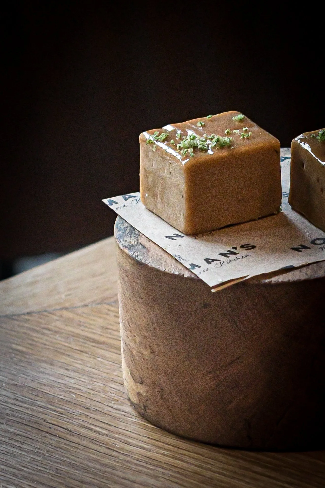 Close-up of two squares of caramel fudge topped with chopped nuts, resting on parchment paper on a wooden surface.