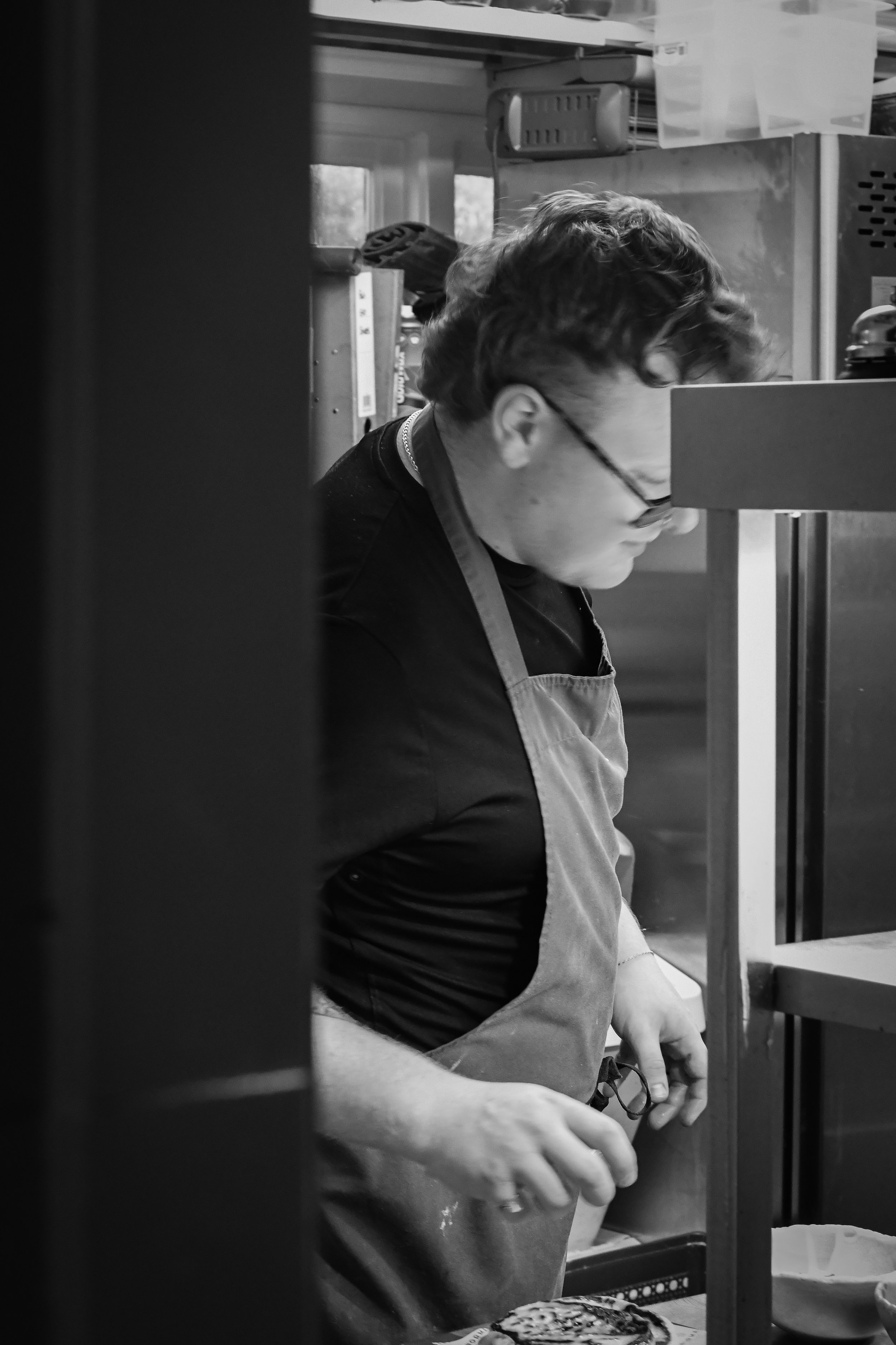 A person with curly hair and glasses, wearing an apron, is working in a kitchen or workshop space. The person is focused on a task, possibly preparing food or working on a craft project.