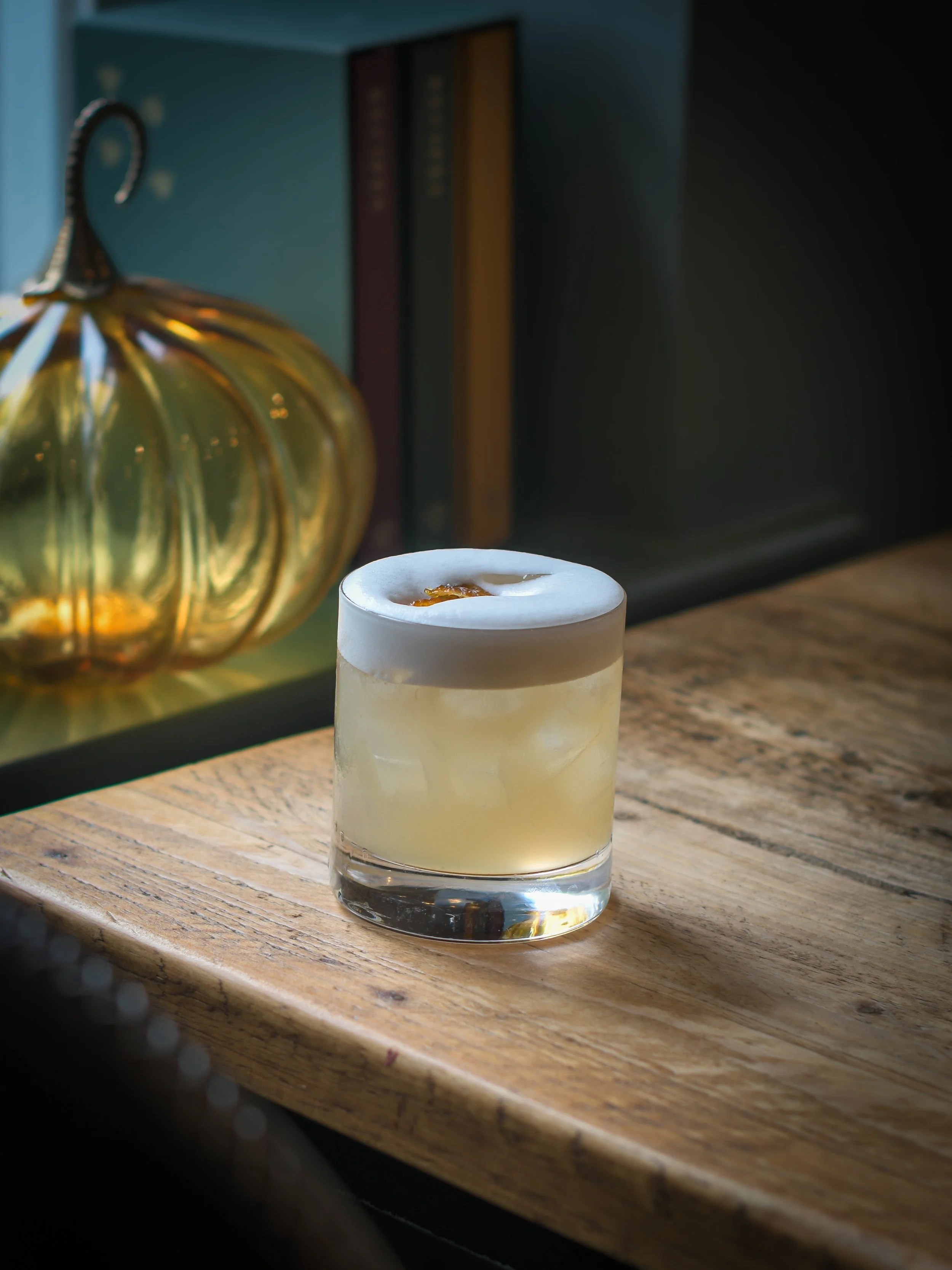 A white candle with a small flame on a wooden surface next to a gold pumpkin decoration and a bookshelf in the background.