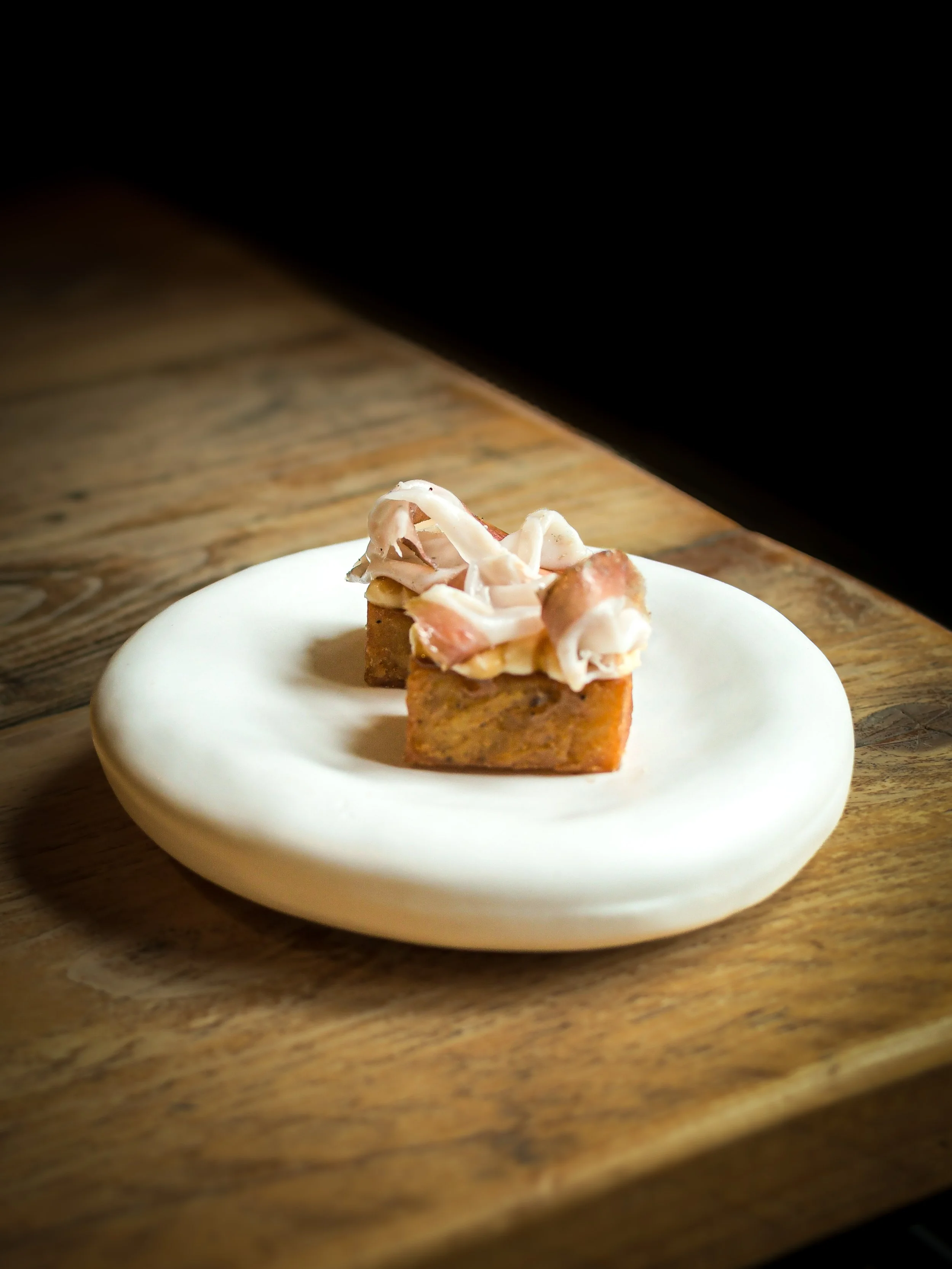 Small square piece of toasted bread topped with slices of ham and cheese, placed on a white round plate on a wooden table.