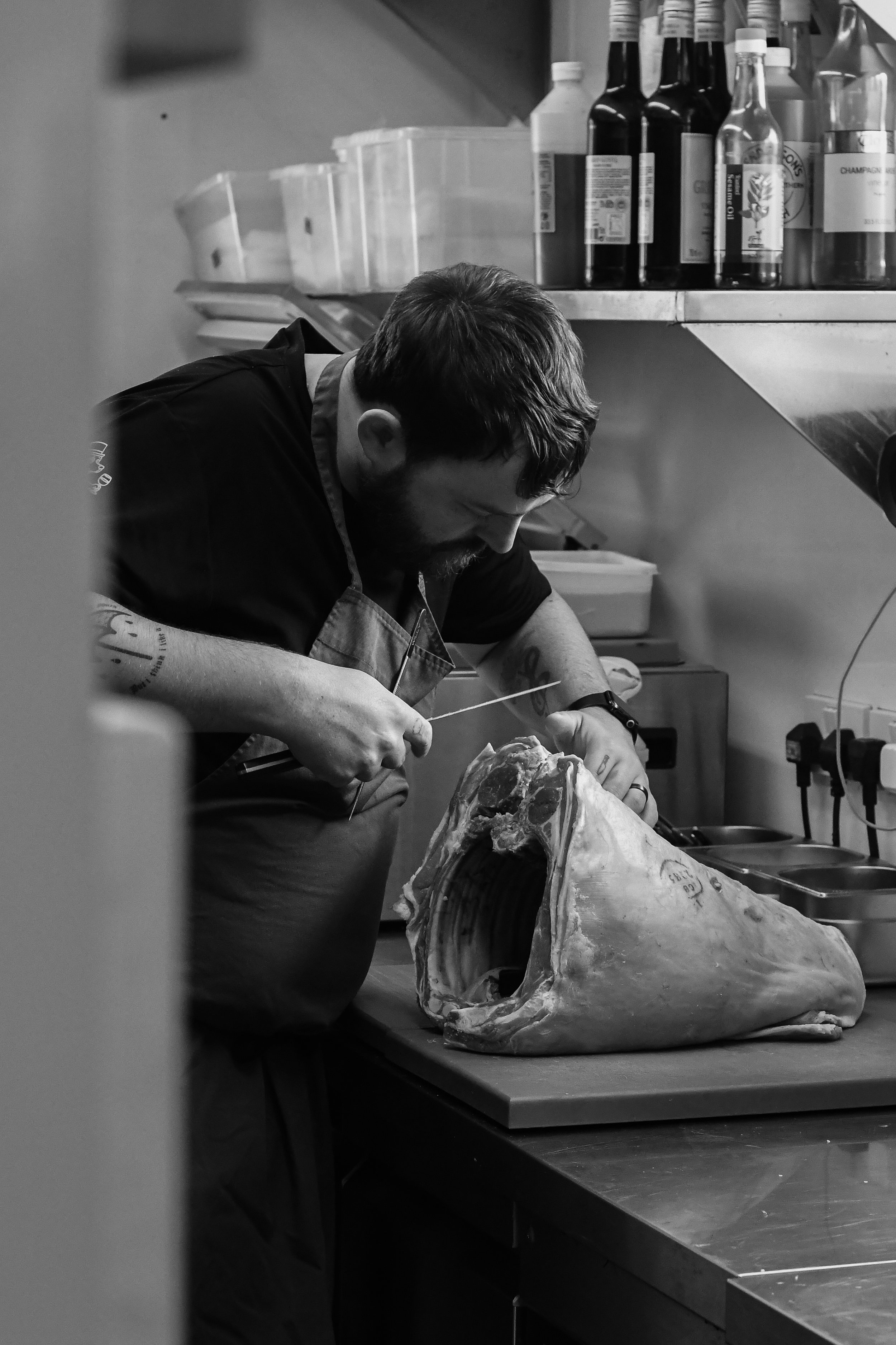 A person in a kitchen working on a large animal skull, possibly a whale, with bottles and containers on shelves in the background.